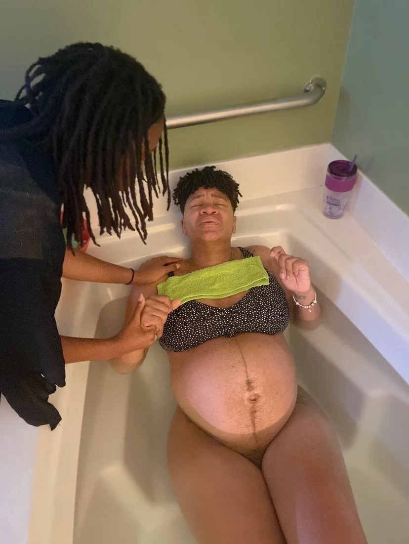 Pregnant person in a bathtub with another person supporting them, holding a green cloth on their chest.