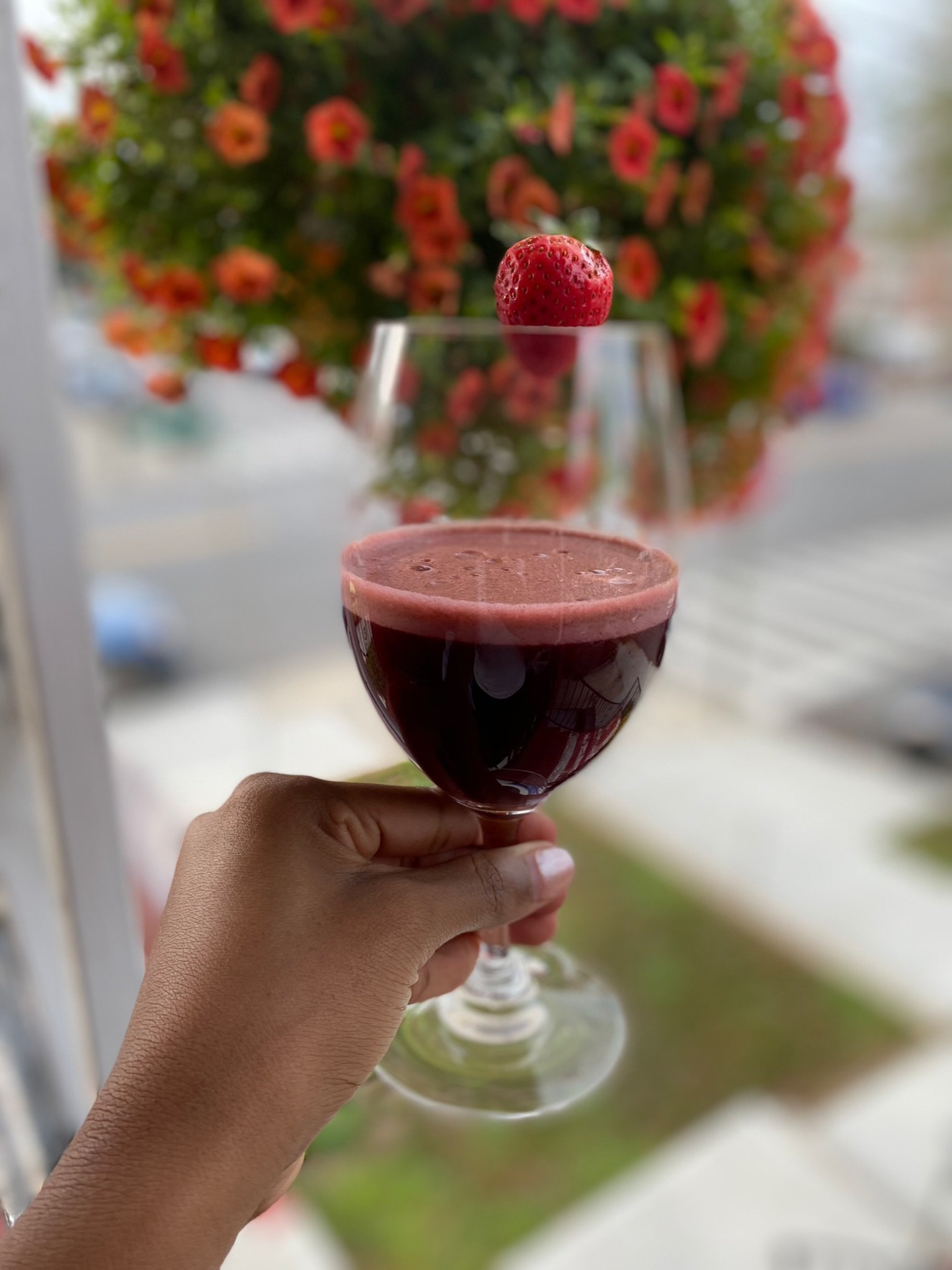 A hand holding a glass of red wine with a strawberry garnish, flowers blurred in the background.