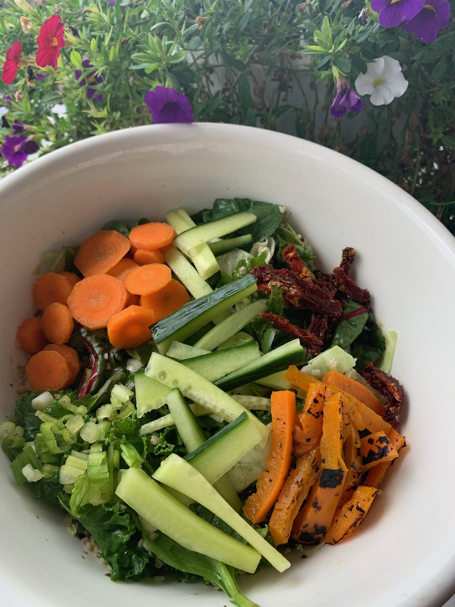 Bowl of fresh salad with ingredients like sliced carrots, cucumbers, bell peppers, lettuce, and sundried tomatoes, placed against a background of vibrant flowers.