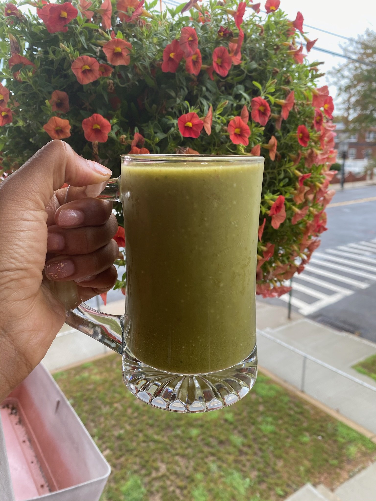 Person holding a glass mug filled with green smoothie, in front of hanging red petunia flowers outdoors.