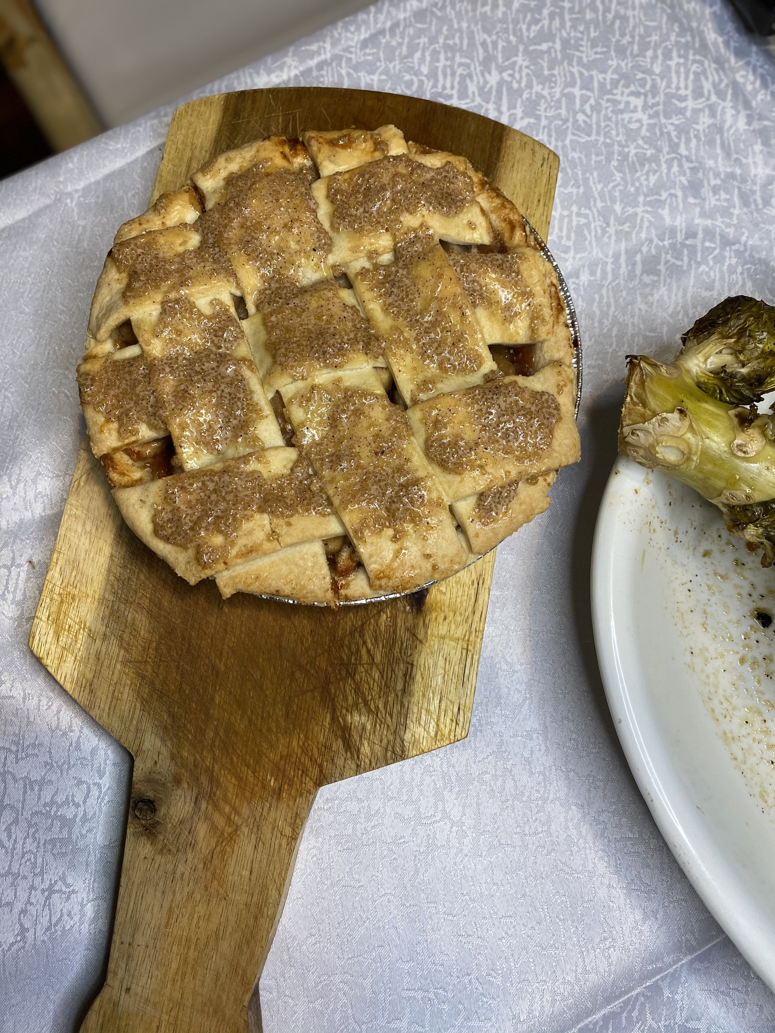 Lattice-topped apple pie on wooden board next to cooked broccoli on white plate.