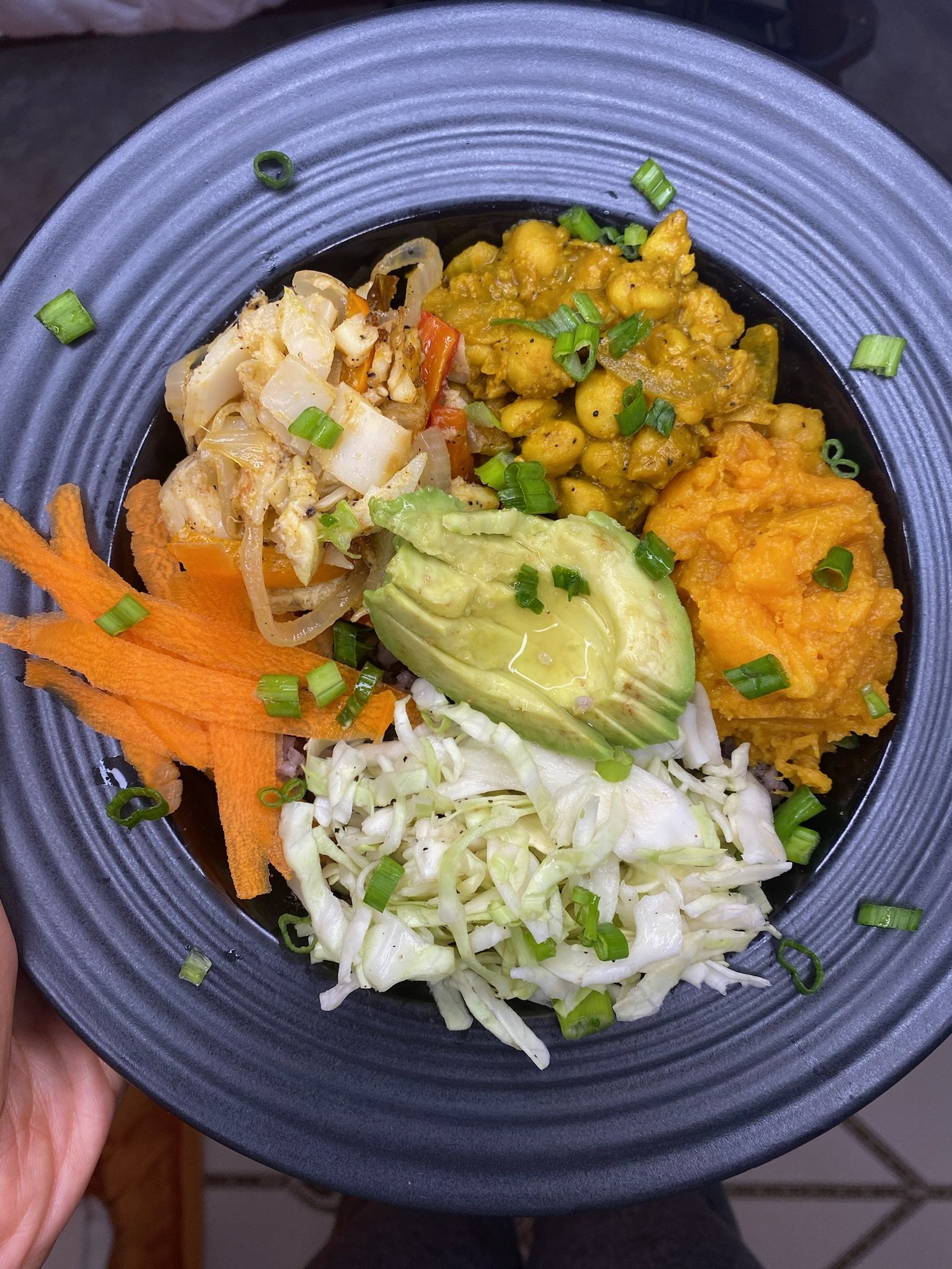 Bowl containing chickpea curry, mashed sweet potatoes, sliced avocado, sautéed vegetables, cabbage slaw, and carrot shavings, garnished with green onions.