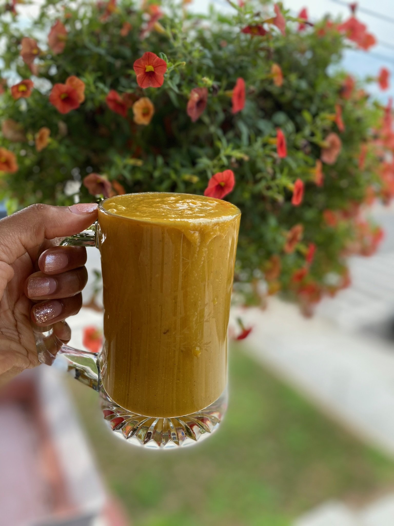 Glass mug filled with yellow smoothie held in hand, blurred background with red flowers and greenery.