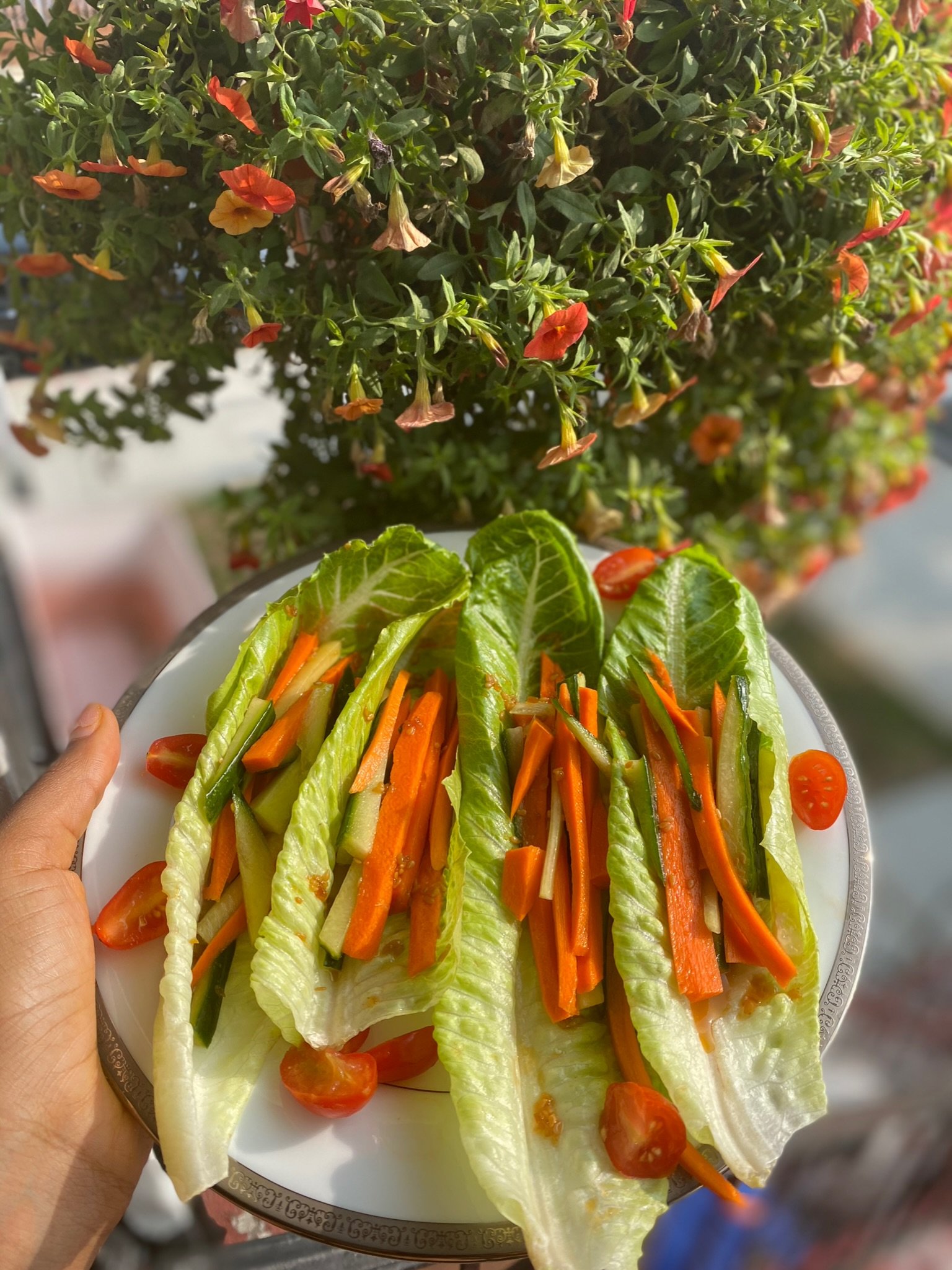 Plate of lettuce wraps with carrots, cucumbers, and cherry tomatoes, held next to red and orange flowers.
