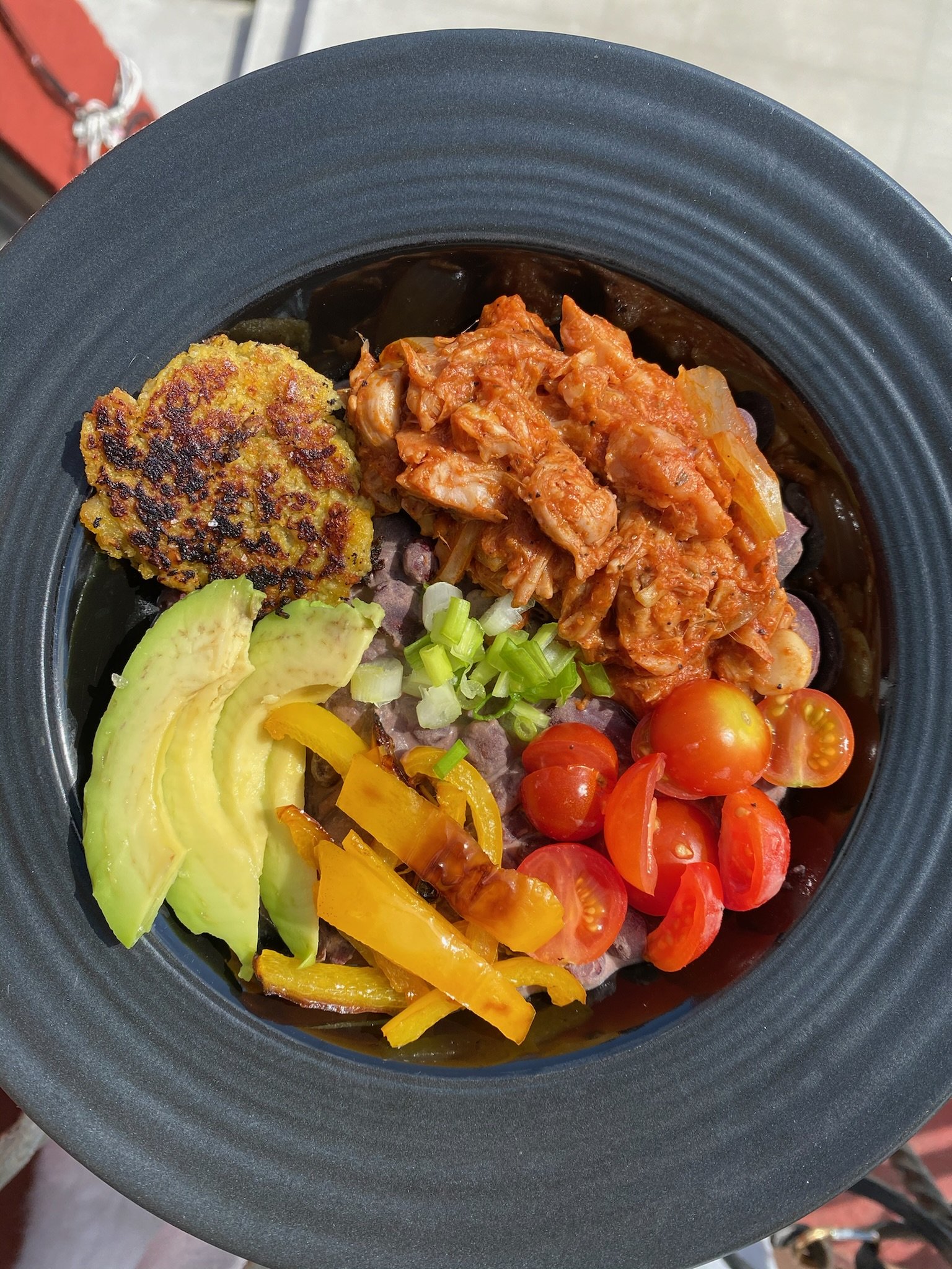 A bowl with sliced avocado, cherry tomatoes, shredded kimchi, scallions, yellow bell peppers, black beans, and a small patty on a black plate.