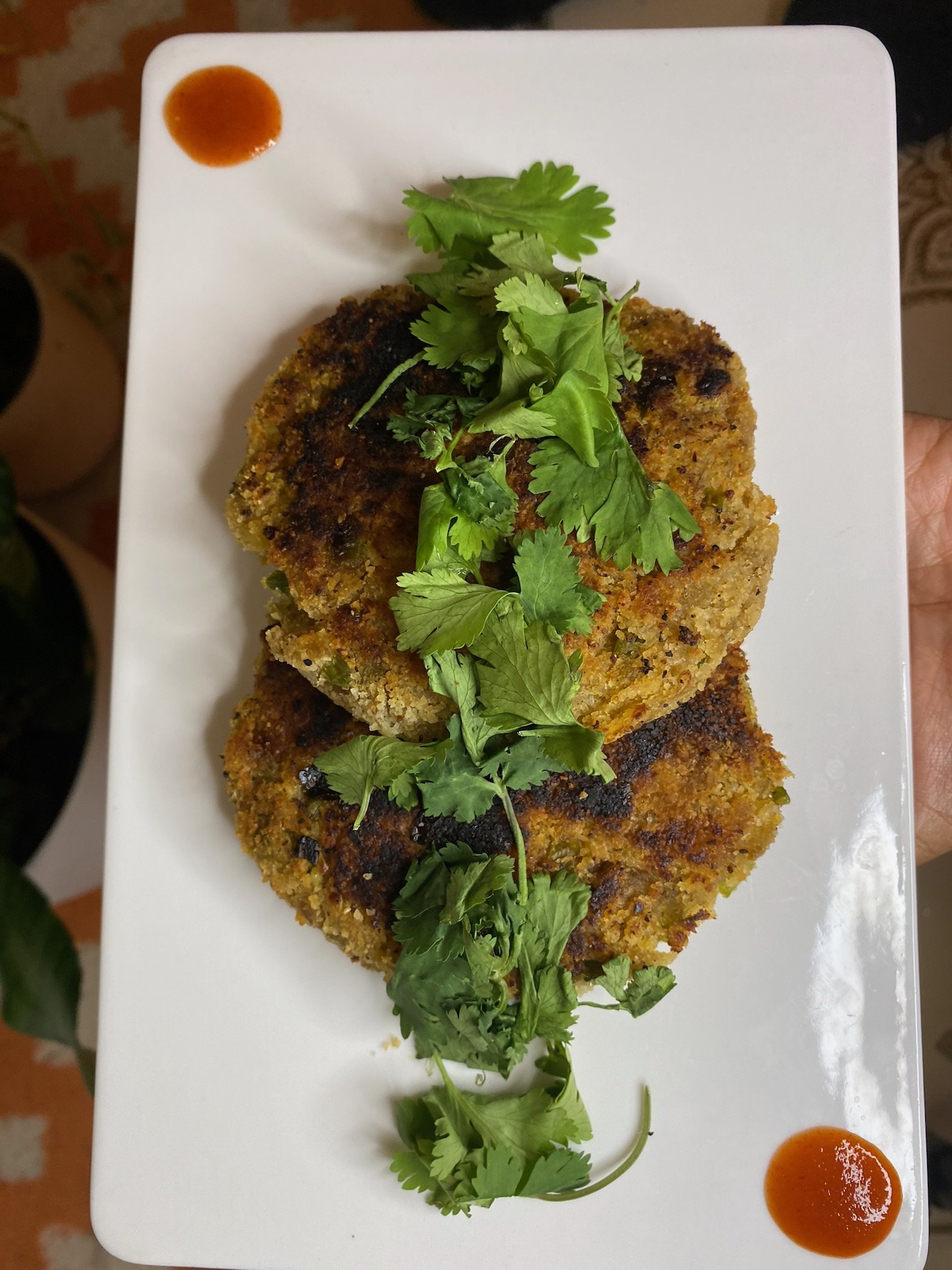 Two round savory patties garnished with fresh cilantro on a white rectangular plate, accompanied by small dollops of red sauce at the corners.