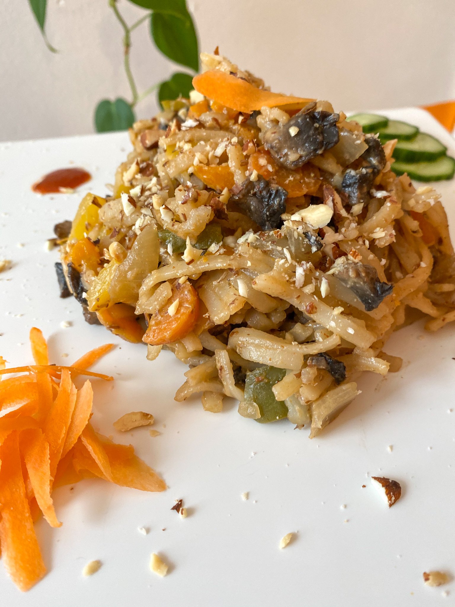 Close-up of a plate with vegetable stir-fry, featuring rice noodles mixed with sliced bell peppers, mushrooms, and topped with crushed nuts. A small portion of grated carrots and sliced cucumbers are arranged decoratively on the side. A leafy plant i