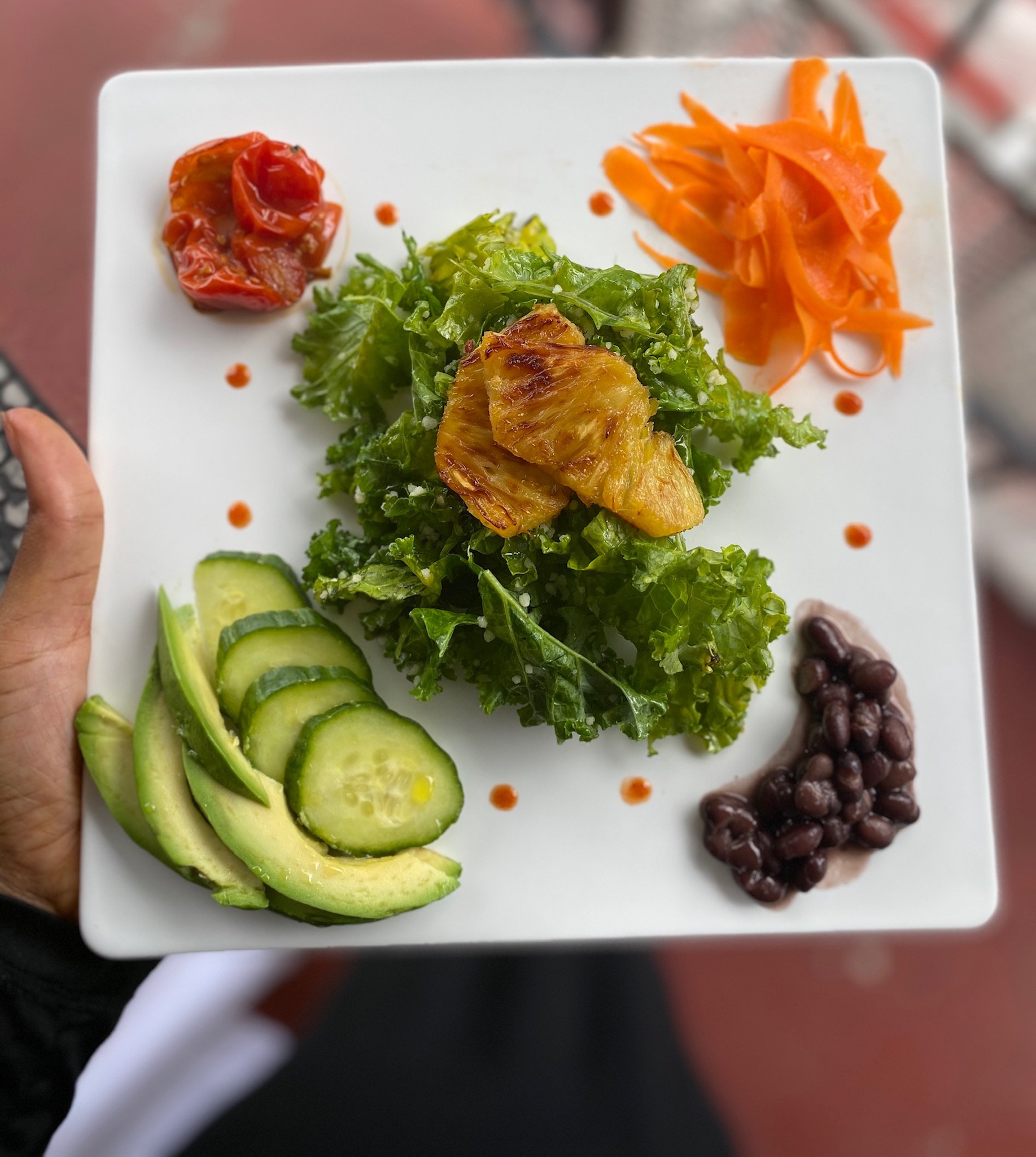 Salad with roasted pineapple, kale, carrot strips, cherry tomatoes, cucumber slices, avocado, and black beans on a white plate.