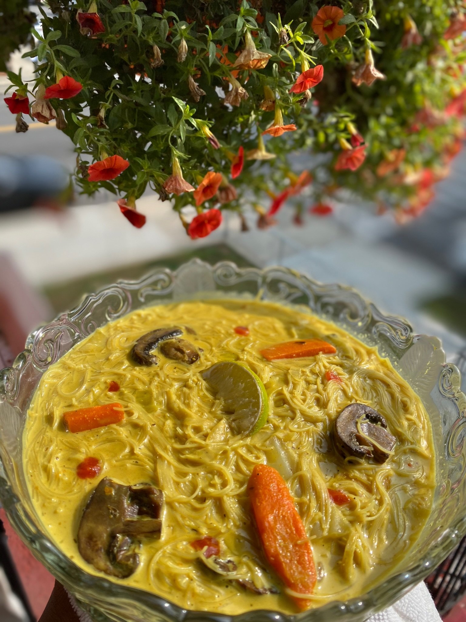 Bowl of yellow curry noodle soup with carrots, mushrooms, and lime slice, under hanging red flowers.