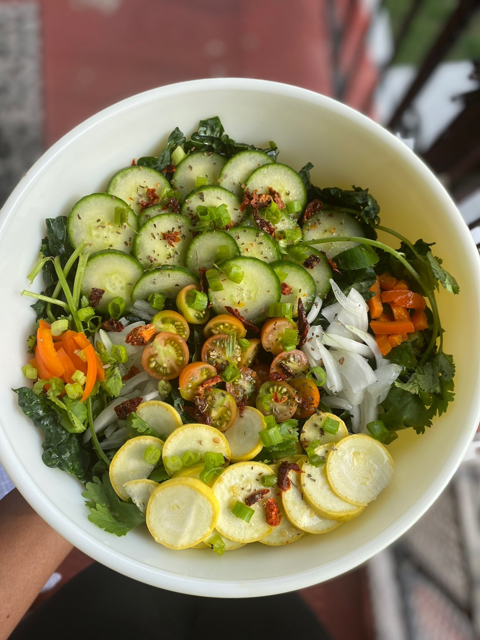 A fresh salad in a white bowl containing sliced cucumbers, cherry tomatoes, yellow squash, and bell peppers. The salad is garnished with green onions and herbs, with a leafy green base.