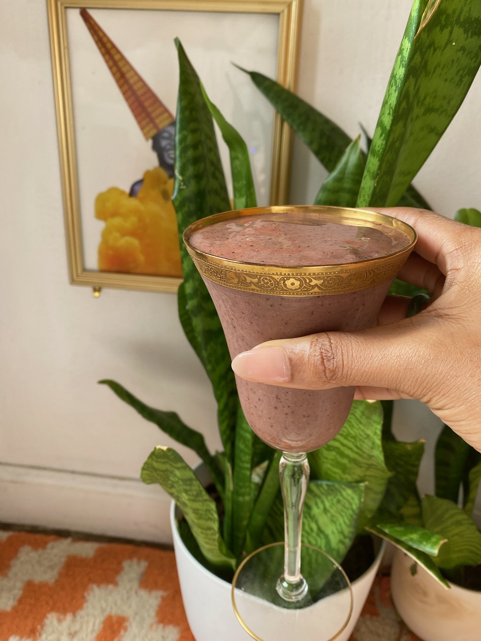 Hand holding a gold-rimmed, purple smoothie in a glass, with indoor plants and a colorful artwork in the background.