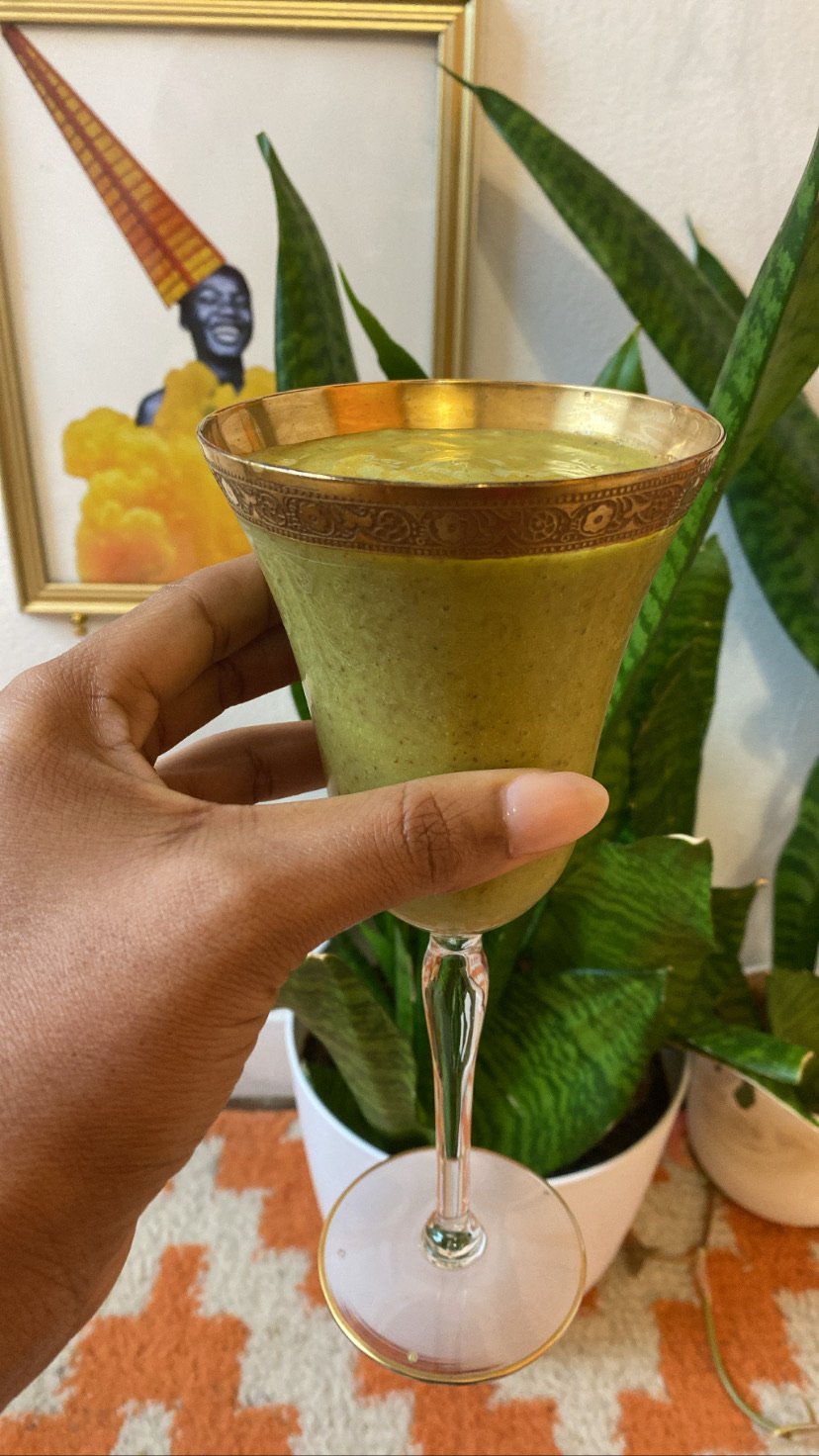 A person holding a glass with a green smoothie in a decorative stemmed glass, in the foreground of a framed picture and a potted plant. The background includes a patterned rug.