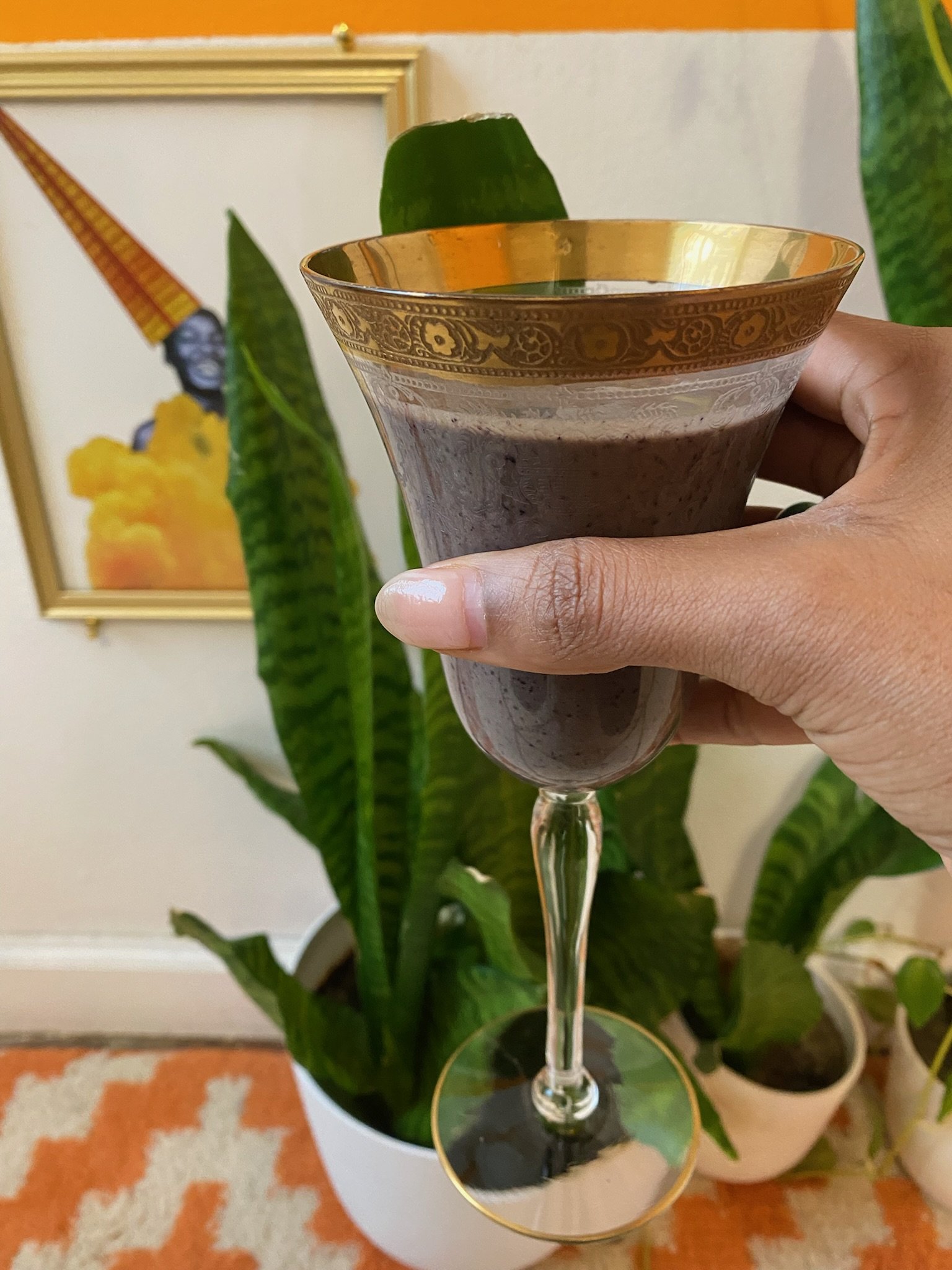 A hand holding a decorative glass with a gold rim containing a dark-colored beverage. In the background, there are houseplants and a framed artwork.