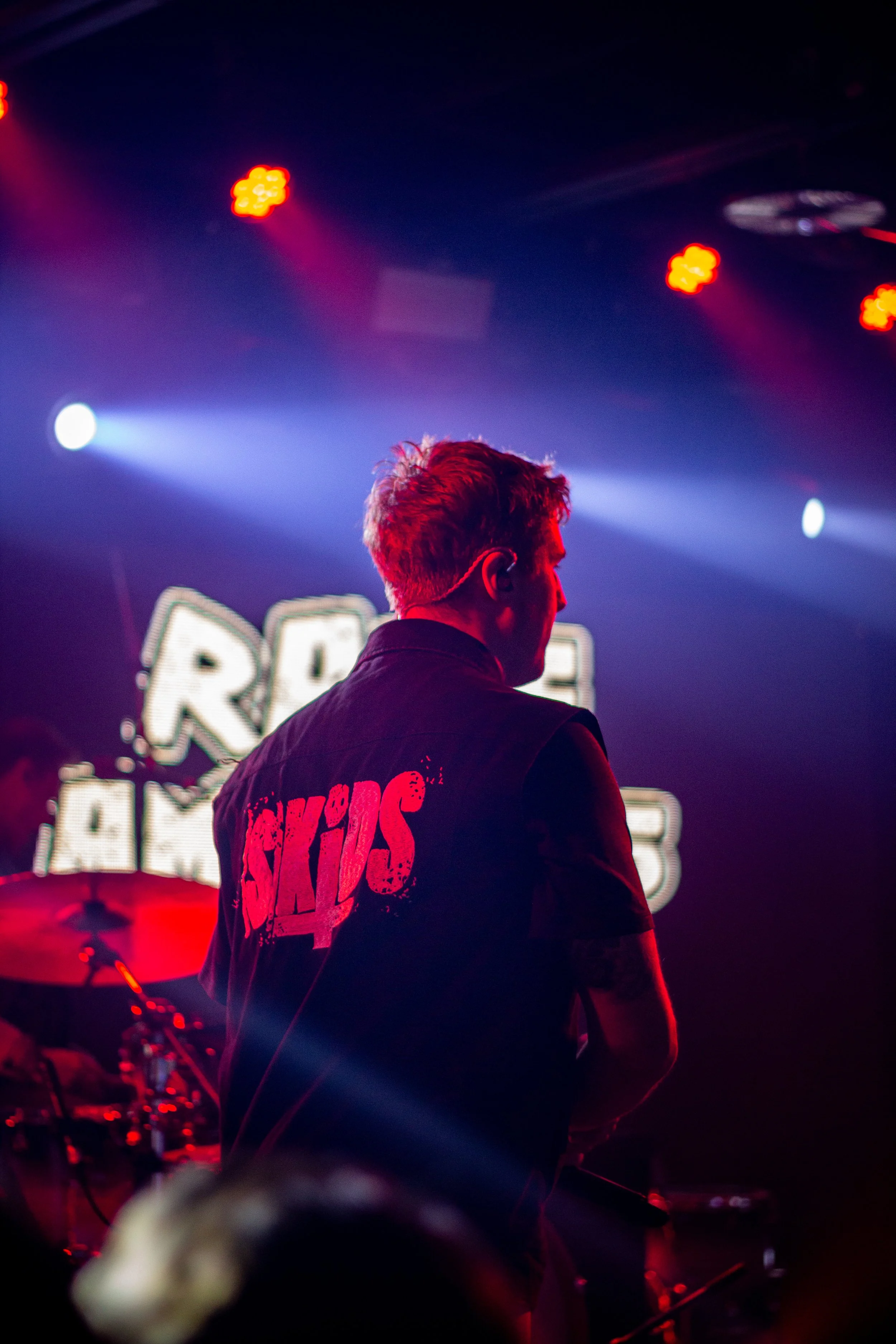 A person with short hair wearing a black vest with red lettering, standing in front of a stage with colorful lights and a drum set.
