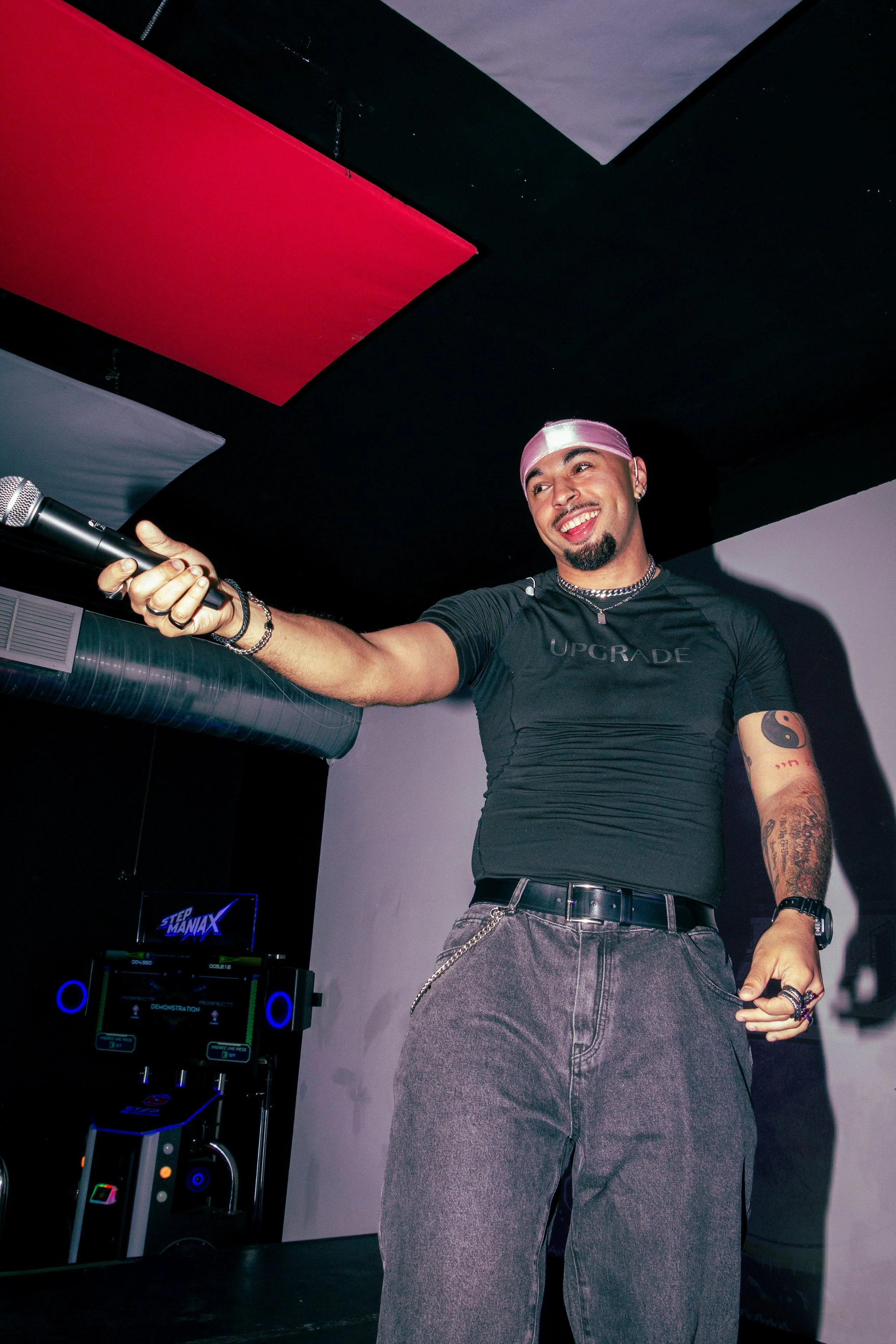 A smiling man wearing a pink headscarf, black t-shirt with 'UPGRADE' written on it, gray pants with a chain, holding a microphone, standing in front of DJ equipment in a dark room with red and gray ceiling panels.