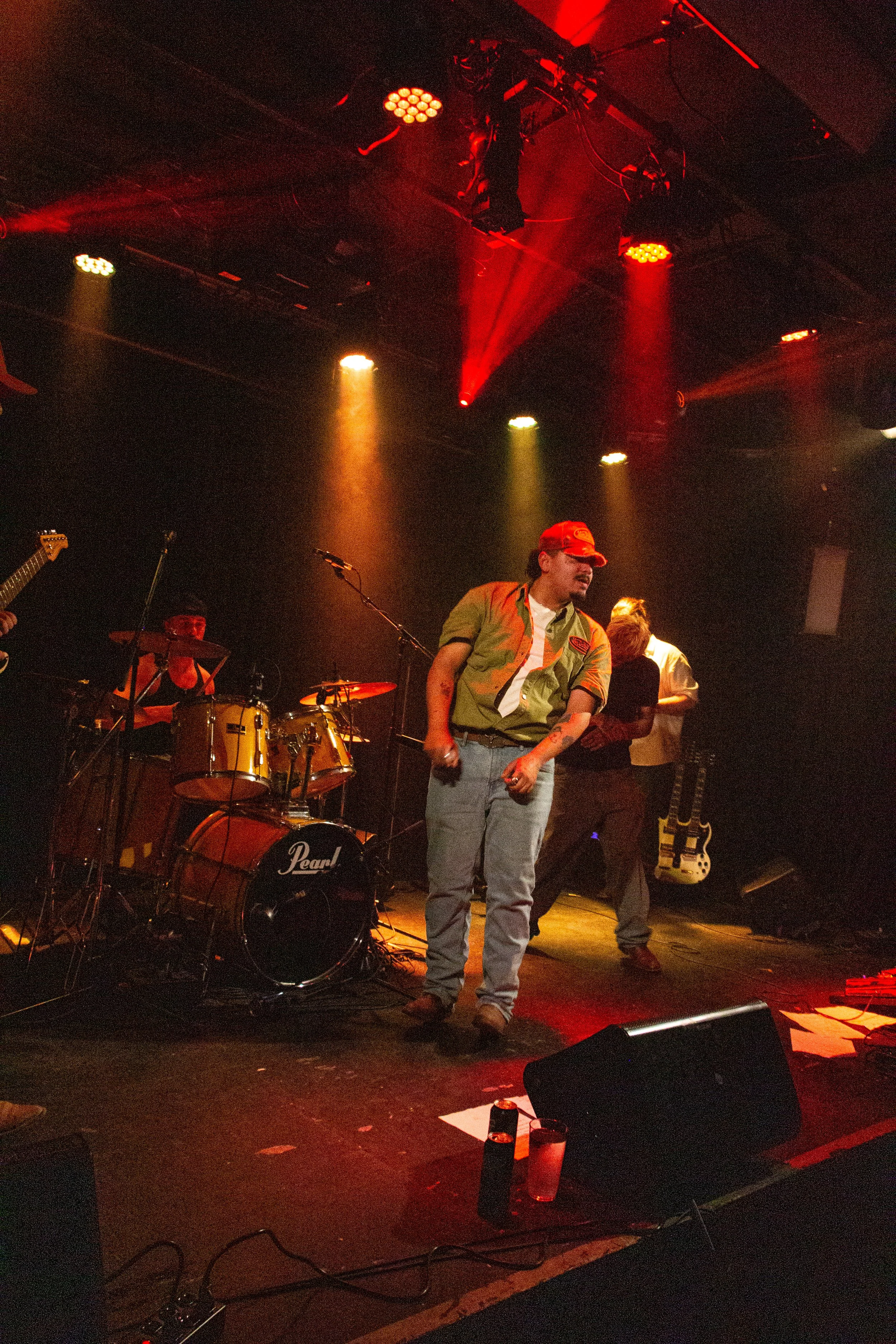 A band performs on stage under red and yellow stage lighting with a singer in a green shirt and red cap, and drums and guitars visible in the background.