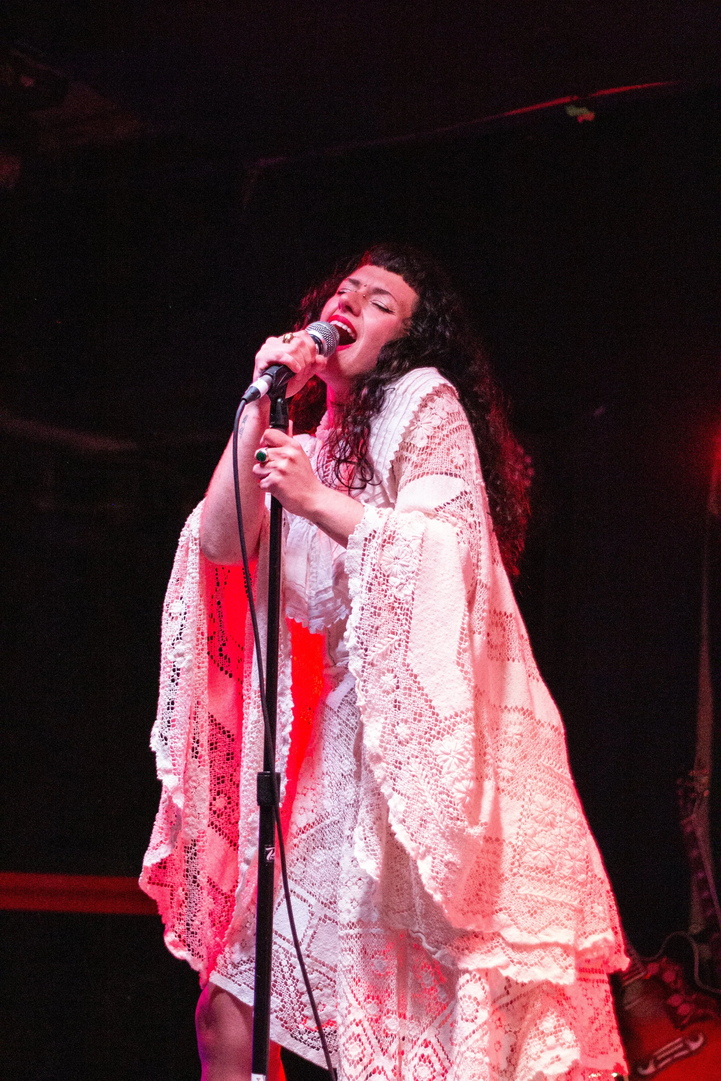 A woman with curly dark hair singing passionately into a microphone on stage, wearing a white lace dress, illuminated by red stage lighting.