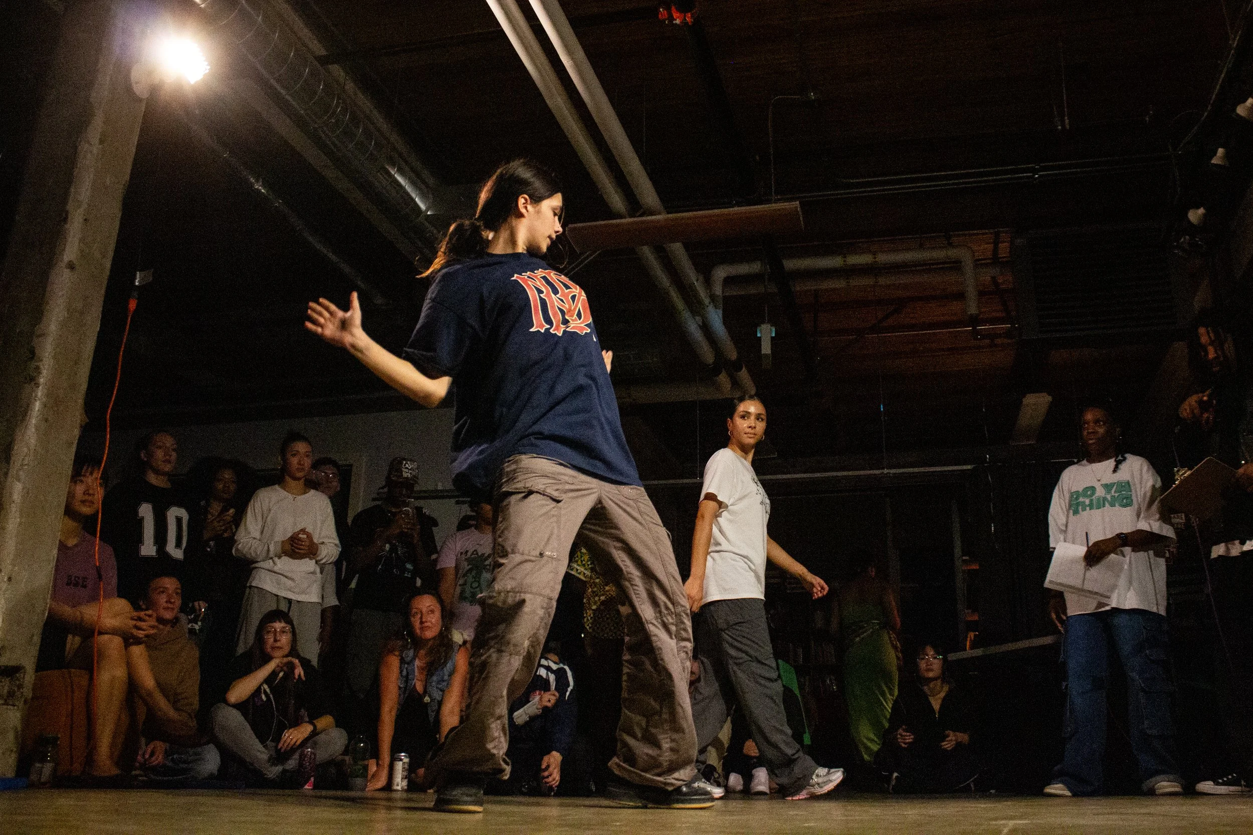 A group of people gathered in a dark indoor space, with some sitting and others standing. In the foreground, a person is dancing or performing, with others watching attentively. The atmosphere appears lively and social.