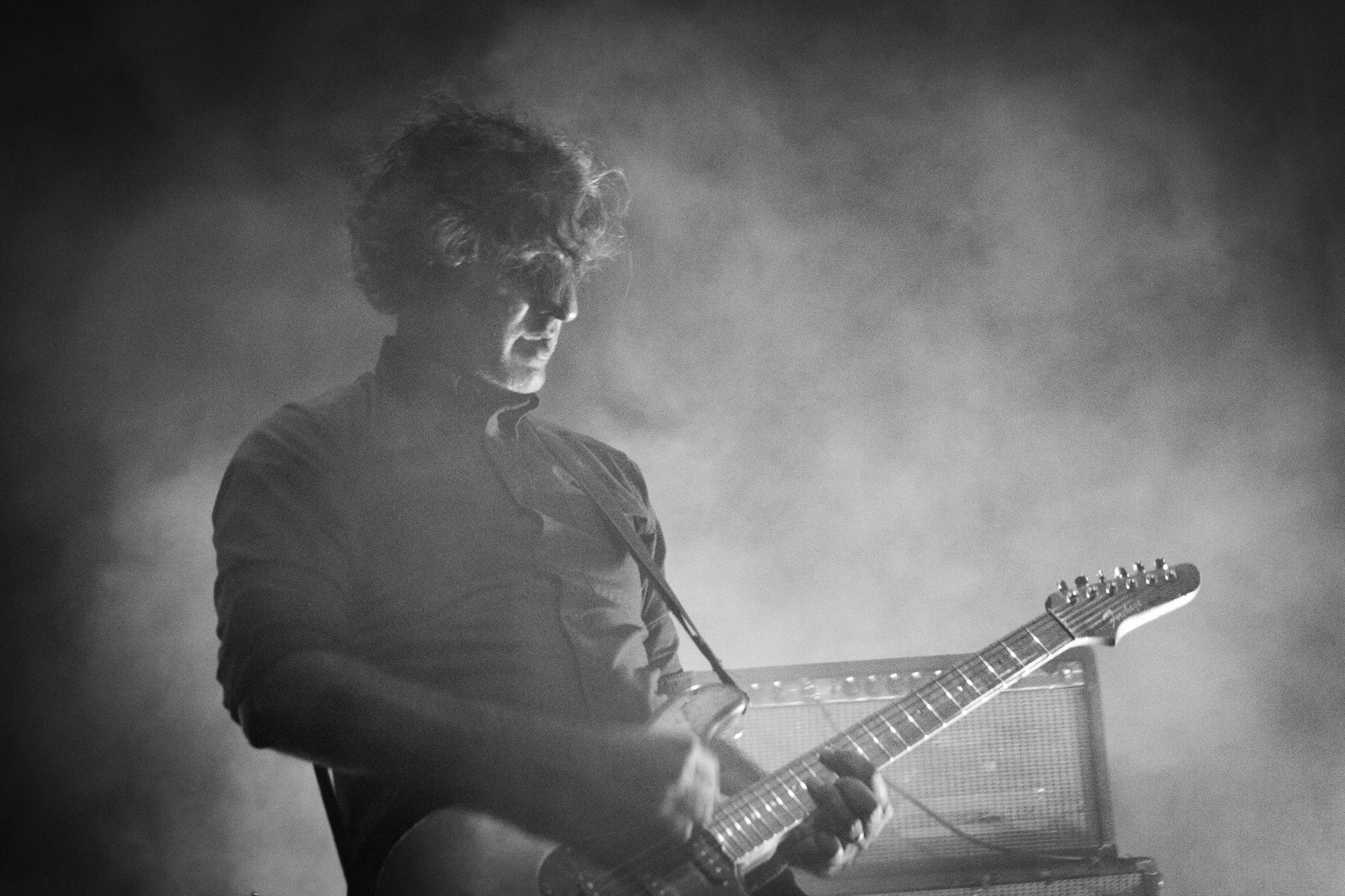 A black and white photo of a man playing an electric guitar, facing left with his eyes closed, wearing glasses and a zip-up jacket, on a dim stage with a misty or smoky atmosphere.