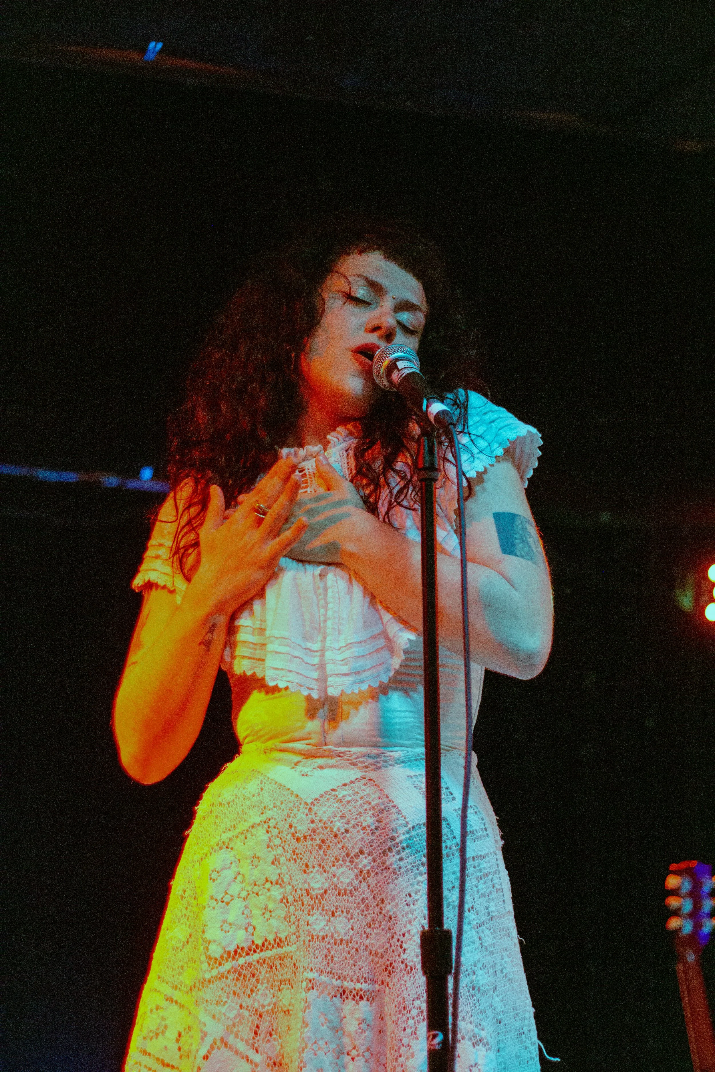 A woman with curly dark hair singing into a microphone, dressed in a white lace dress, with her eyes closed and one hand on her chest in a performance setting with dark background and colorful stage lighting.