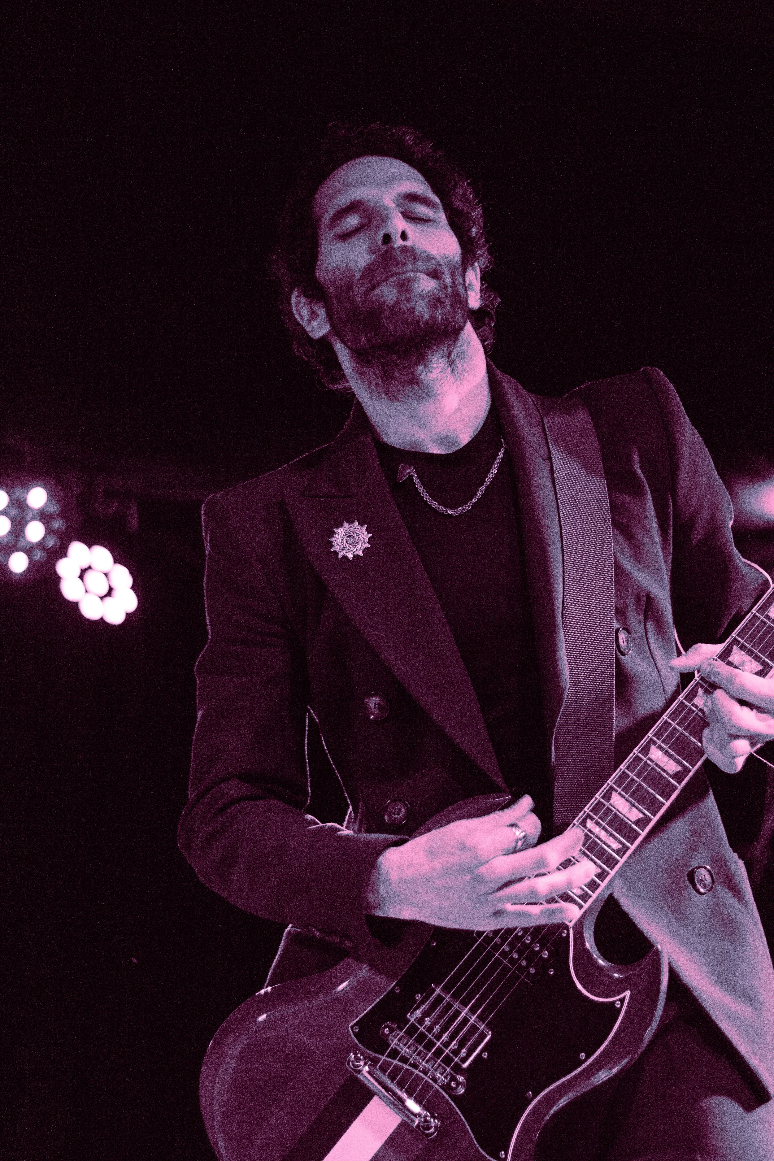 A man with a beard and closed eyes playing an electric guitar on stage, wearing a dark blazer and a chain necklace, with purple stage lighting in the background.
