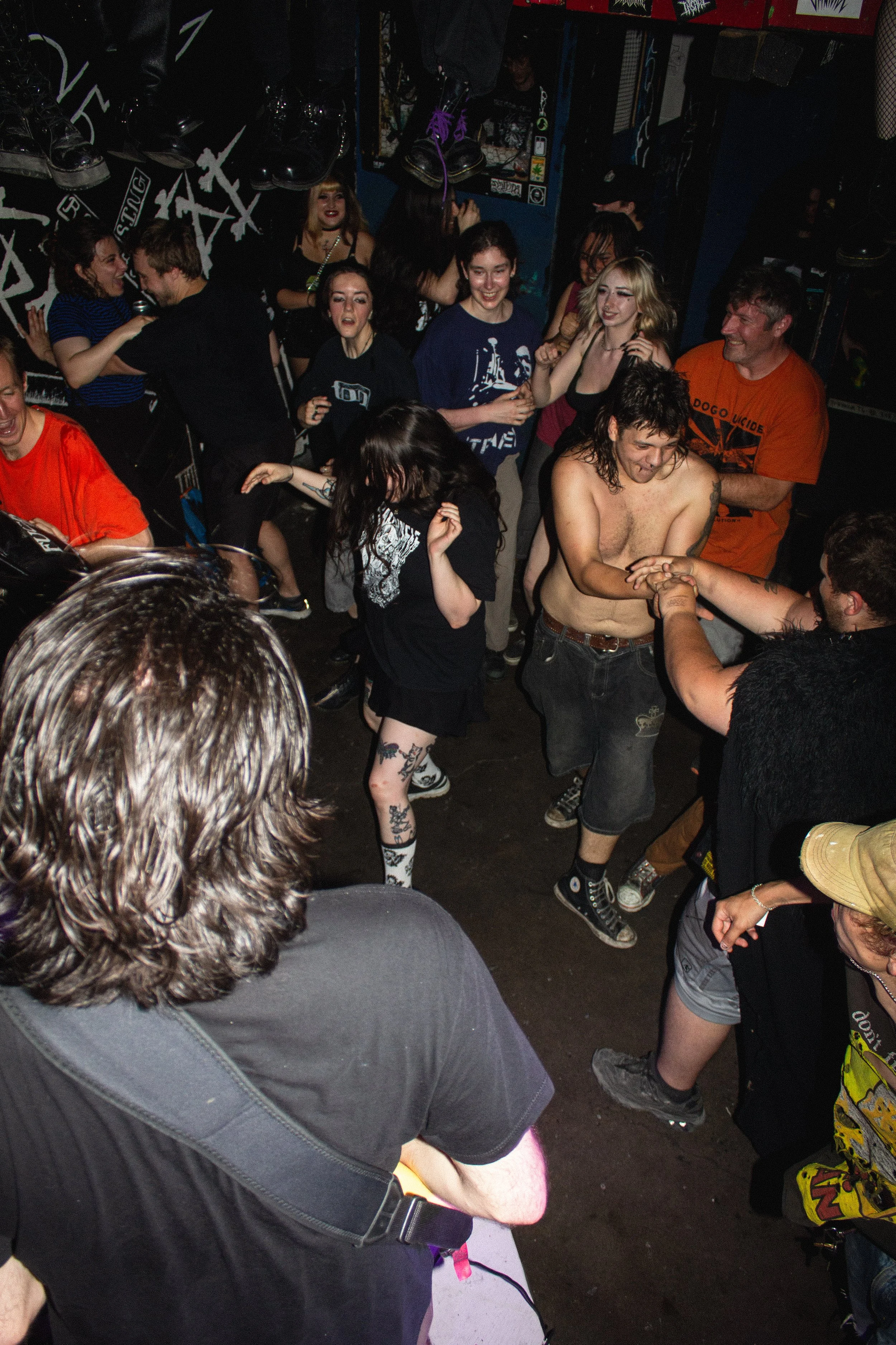 People dancing and socializing in a crowded underground music venue with dark walls and wall art, some with drinks, wearing casual and alternative clothing.