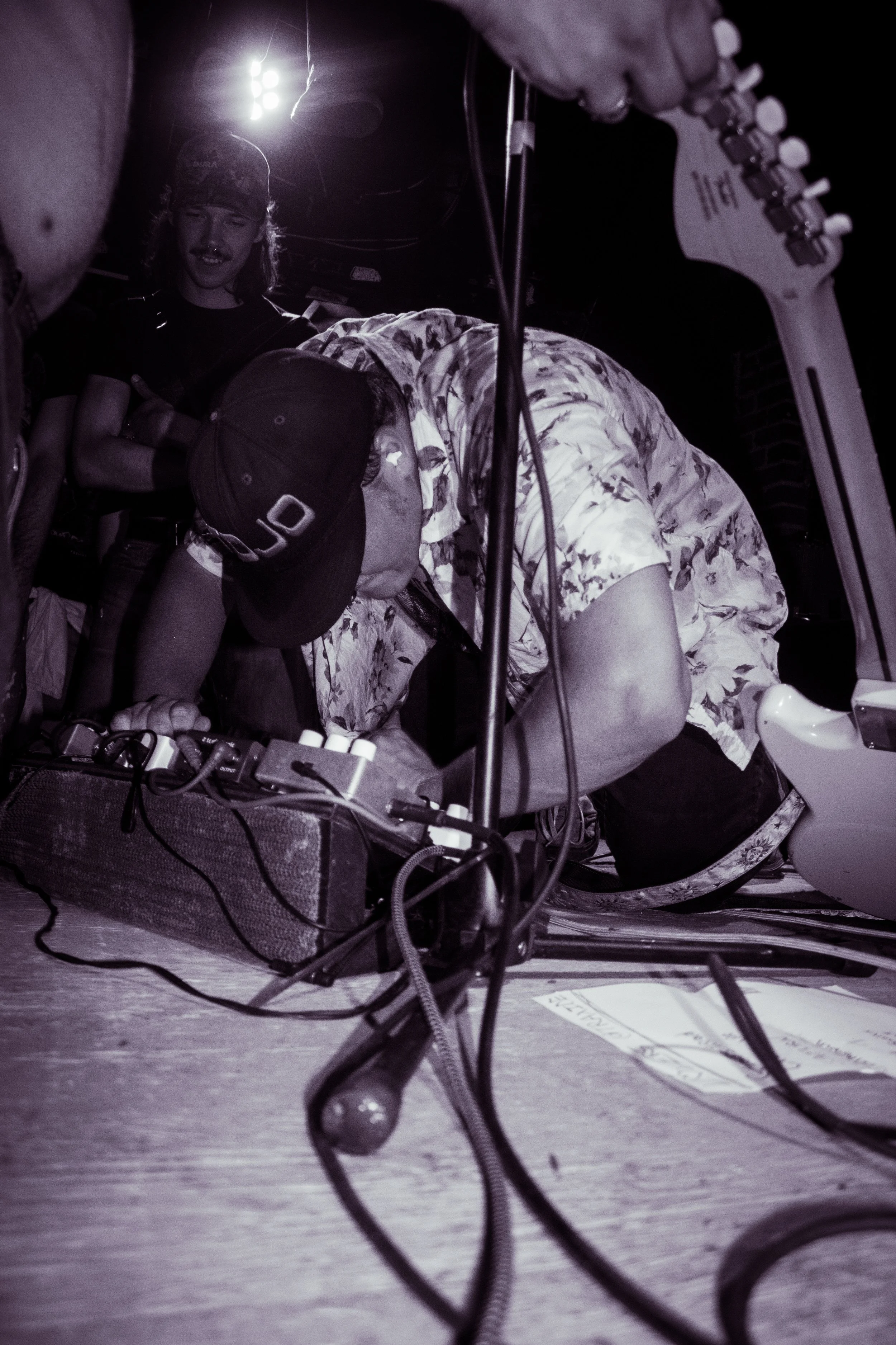 A musician is playing an electric guitar on stage, with a person next to him wearing a black cap and a patterned shirt, possibly mixing music or adjusting equipment. The scene is dark with a bright light source in the background.