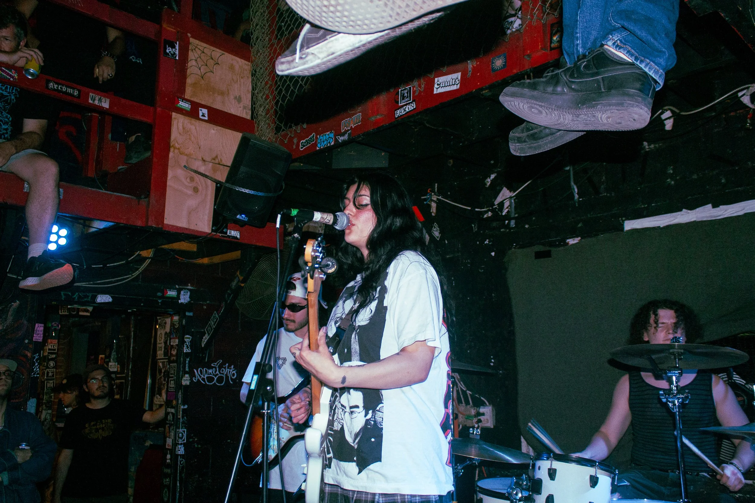 A woman with black hair singing into a microphone while playing an electric guitar at a music venue. Other band members and audience are visible in the background.
