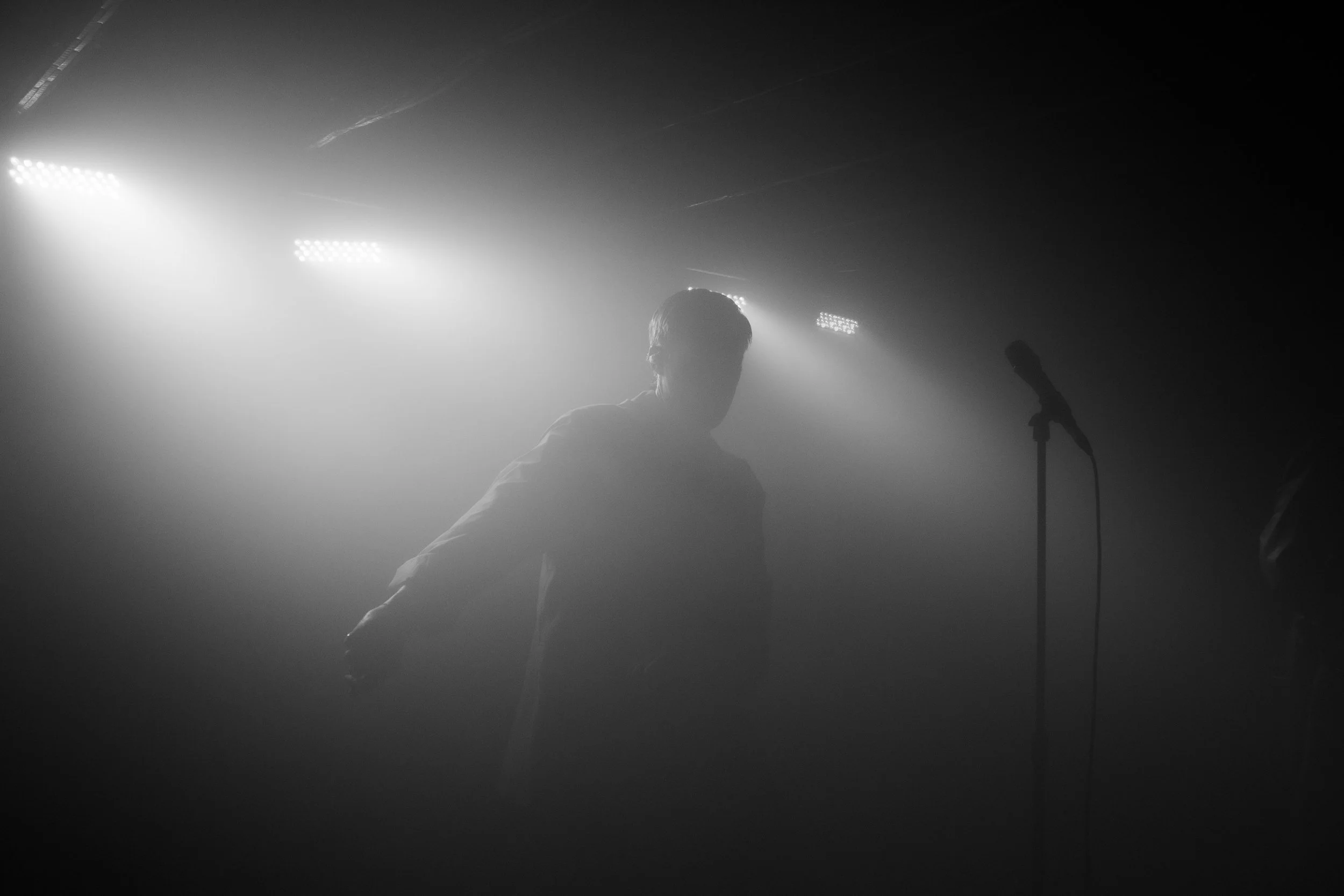A person with glasses is on stage performing with a microphone in a dark, foggy environment, illuminated by bright stage lights.