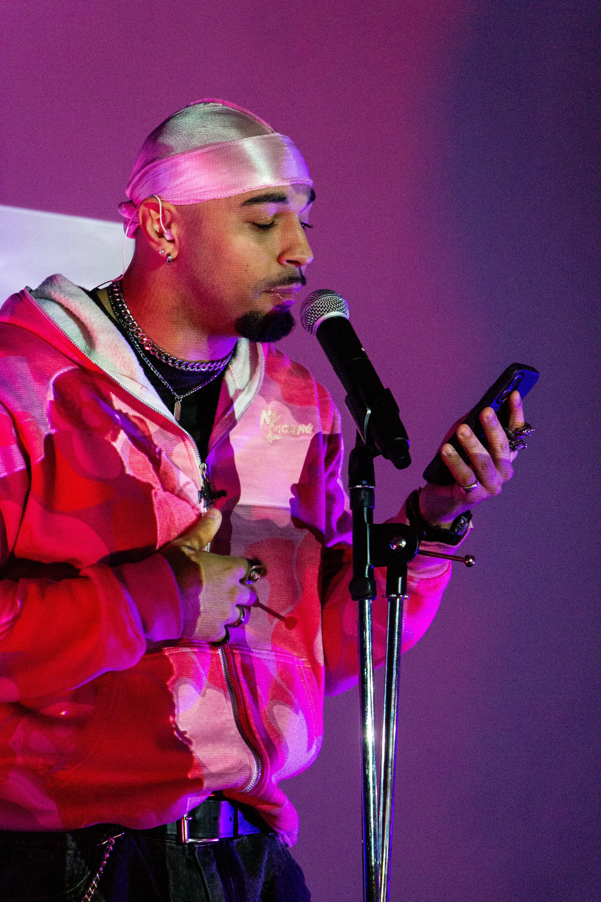 A person wearing a shiny pink head wrap, a pink camouflage jacket, and multiple necklaces, holding a smartphone in front of a microphone, with closed eyes, against a purple-pink background.