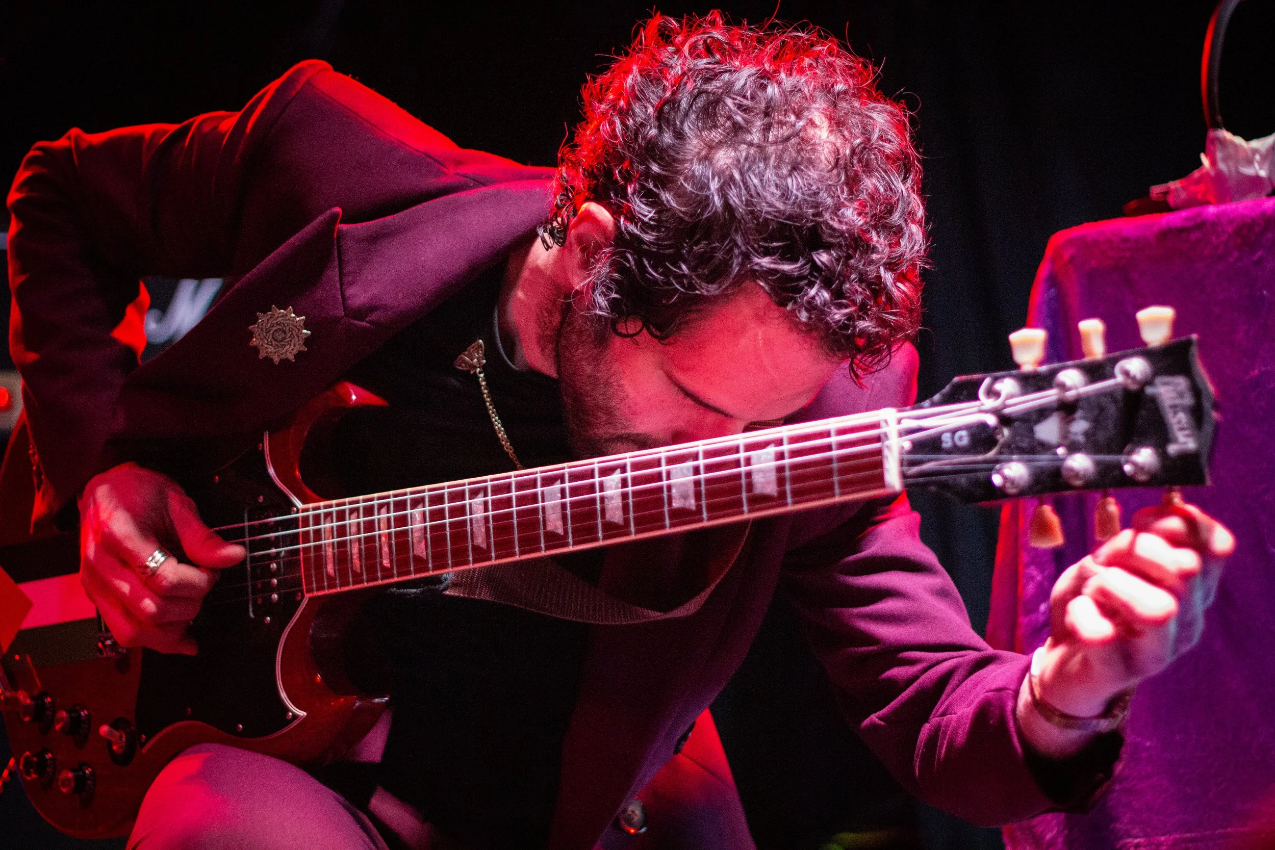 A musician with curly hair plays an electric guitar on stage, illuminated by red and purple lights.