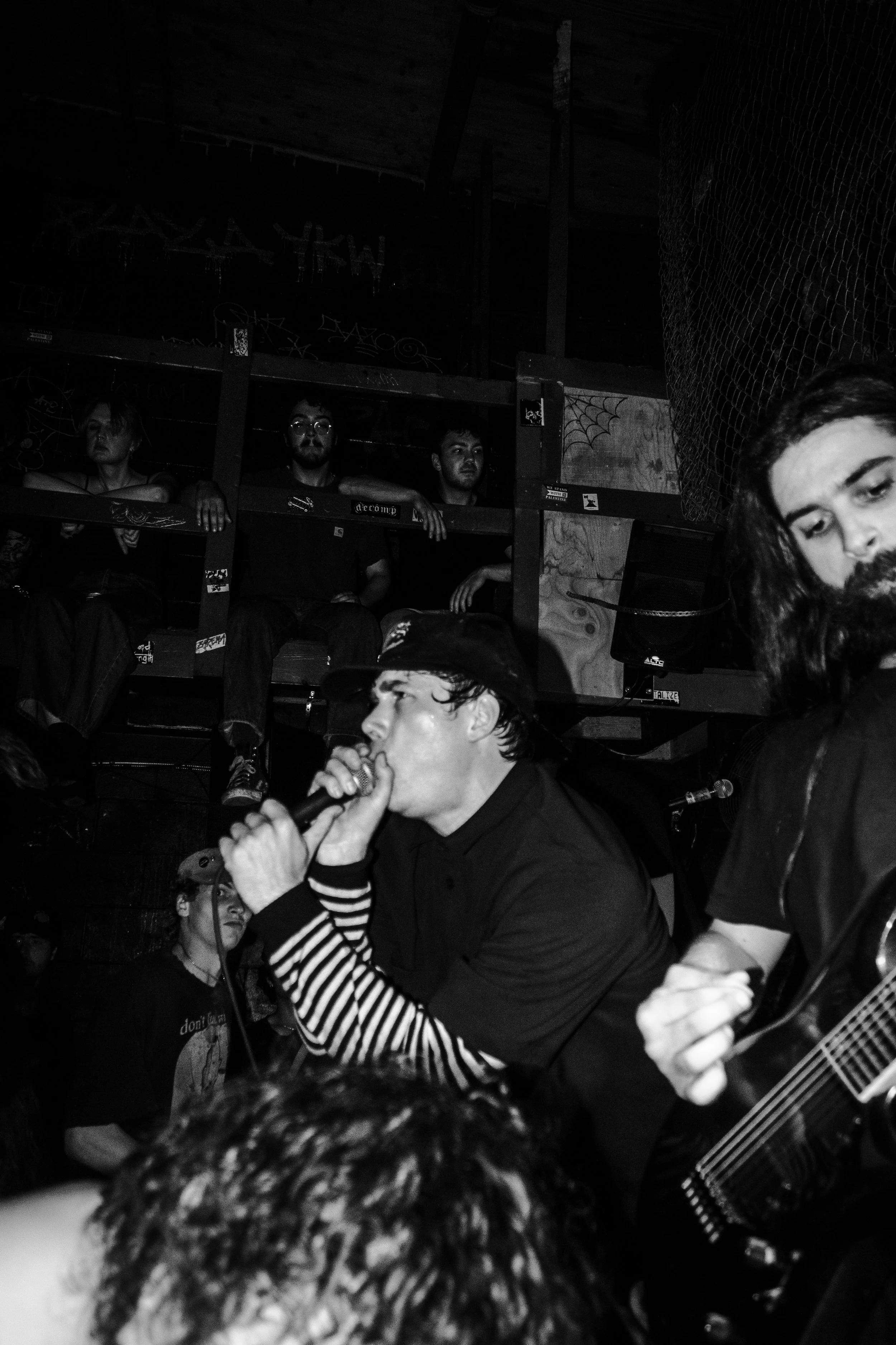 A black and white photo of a group of young adults at a crowded indoor music event. One person in the foreground is singing into a microphone, wearing a striped long-sleeve shirt. Others are sitting or standing behind, some with serious expressions. 