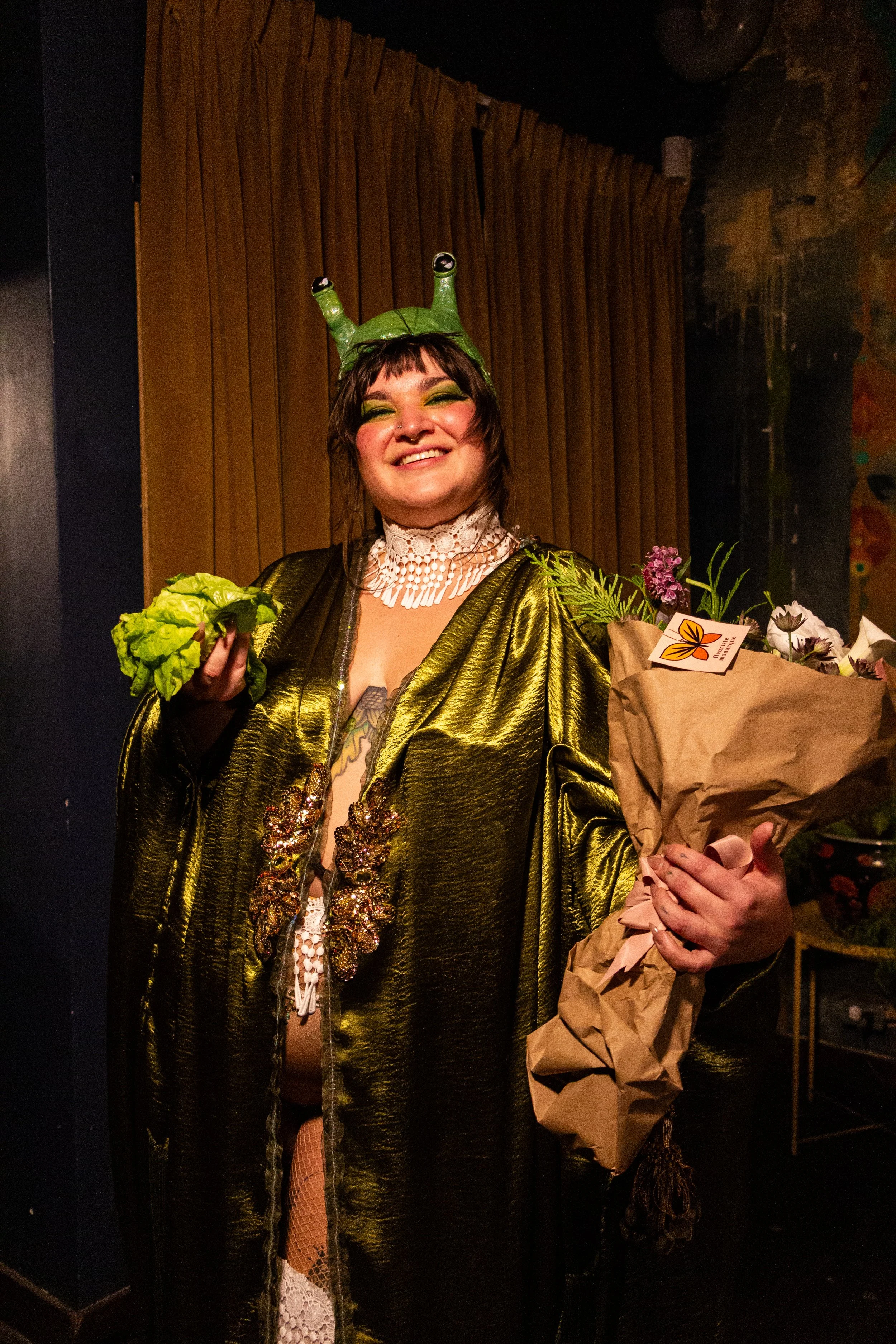 A smiling woman in a shiny gold jacket, green eye makeup, and a frog hat, holding a bouquet of flowers.