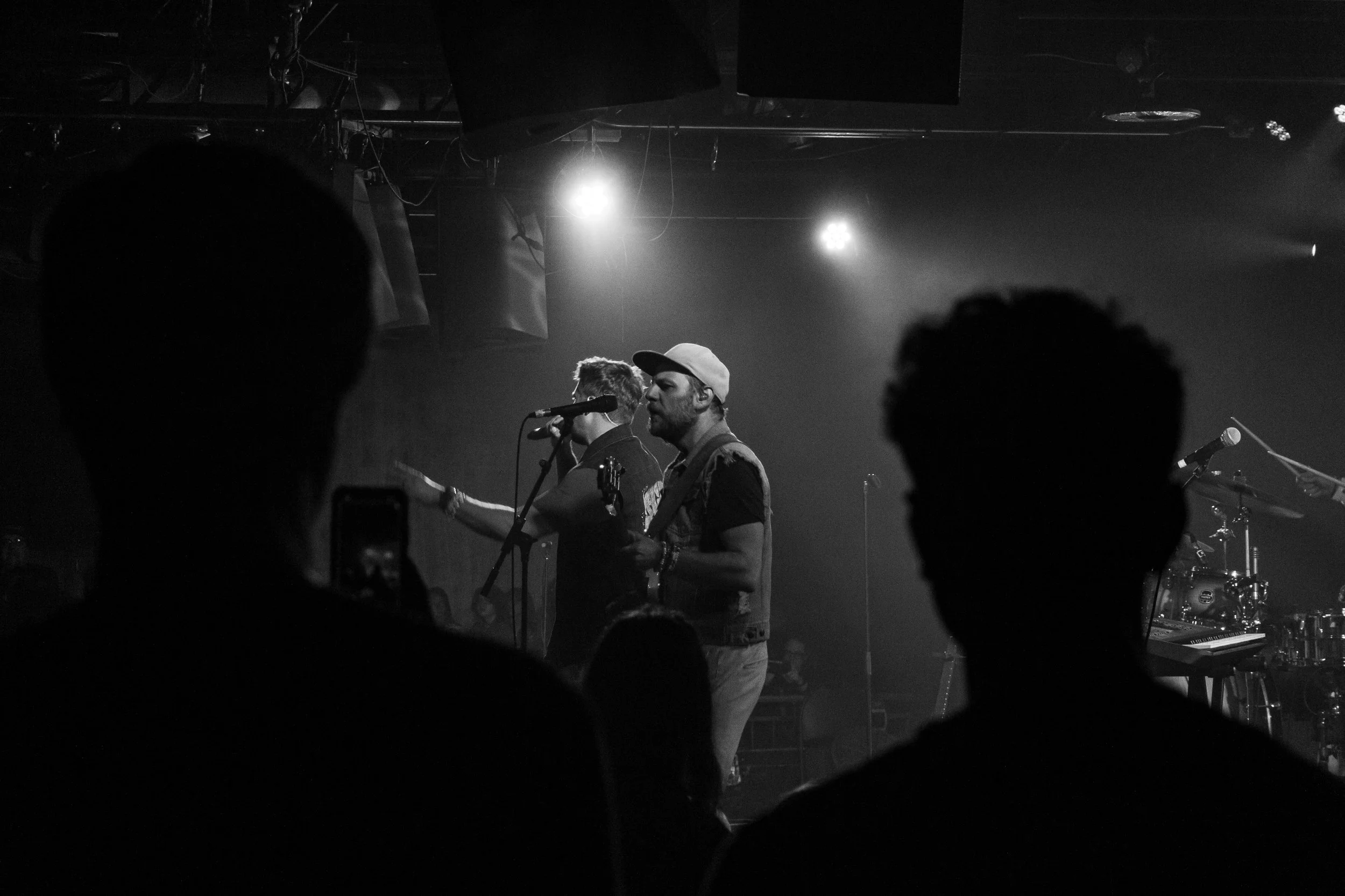 Black and white photo of a live music performance with band members on stage and audience silhouettes in the foreground.
