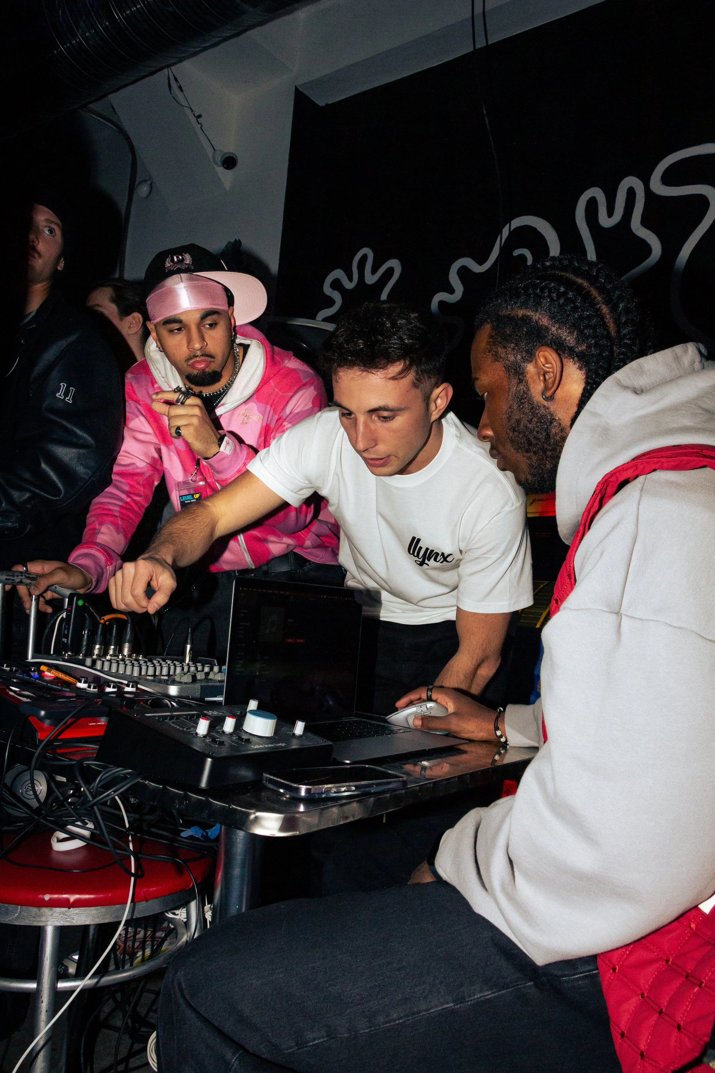 A group of young men in a dimly lit room, focused on a DJ controller, with one holding a game controller. The scene appears to be a casual indoor gathering or DJ session.