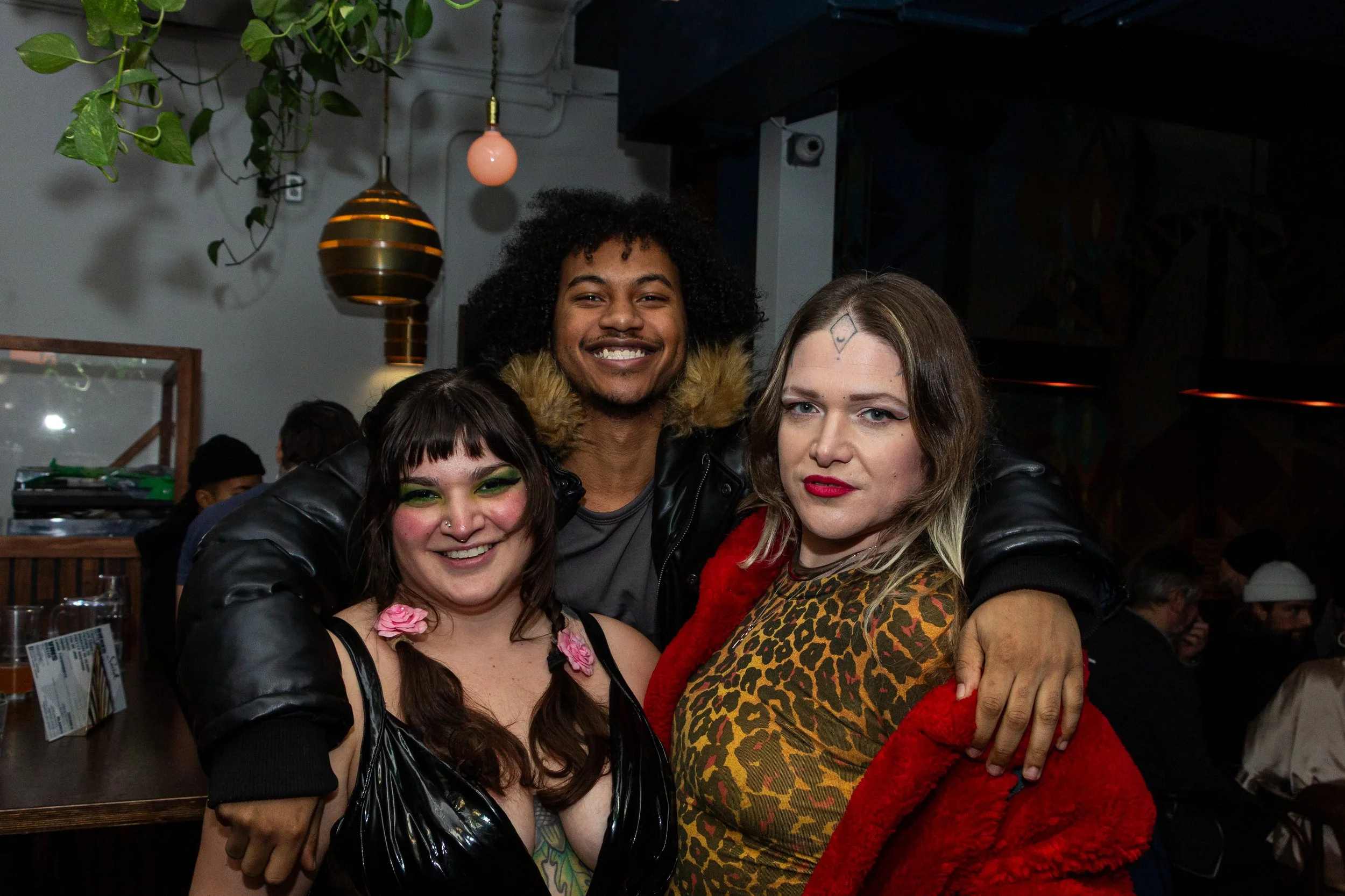 Three women and one man posing together at a social gathering. The woman on the left has dark hair, green eyeshadow, a nose piercing, and is wearing a black outfit with a pink flower accessory near her chest. The man in the middle has curly hair, a b