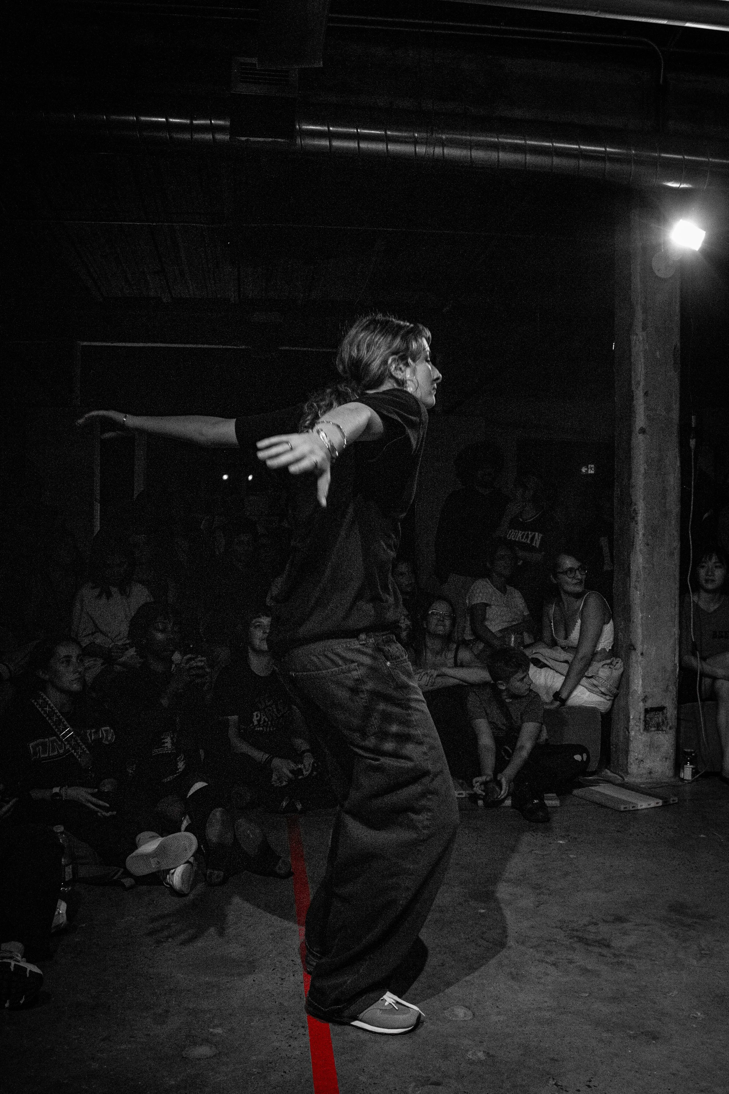 A woman with long hair, wearing a black outfit and sneakers, dancing with her arm extended in front of a seated audience in a dark indoor setting.