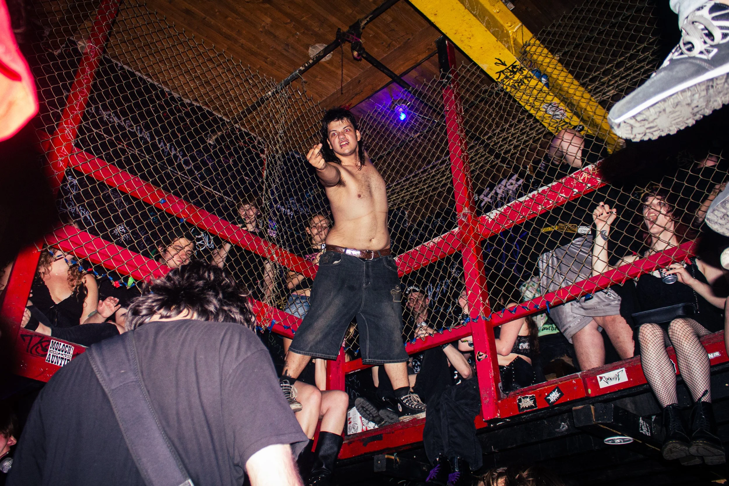 A shirtless man with dark hair and a ring finger tattoo stands in a raised cage area at a concert, making a gesture towards the camera. He is surrounded by people, some of whom are smiling and holding cameras or drinks. The setting is dark with color