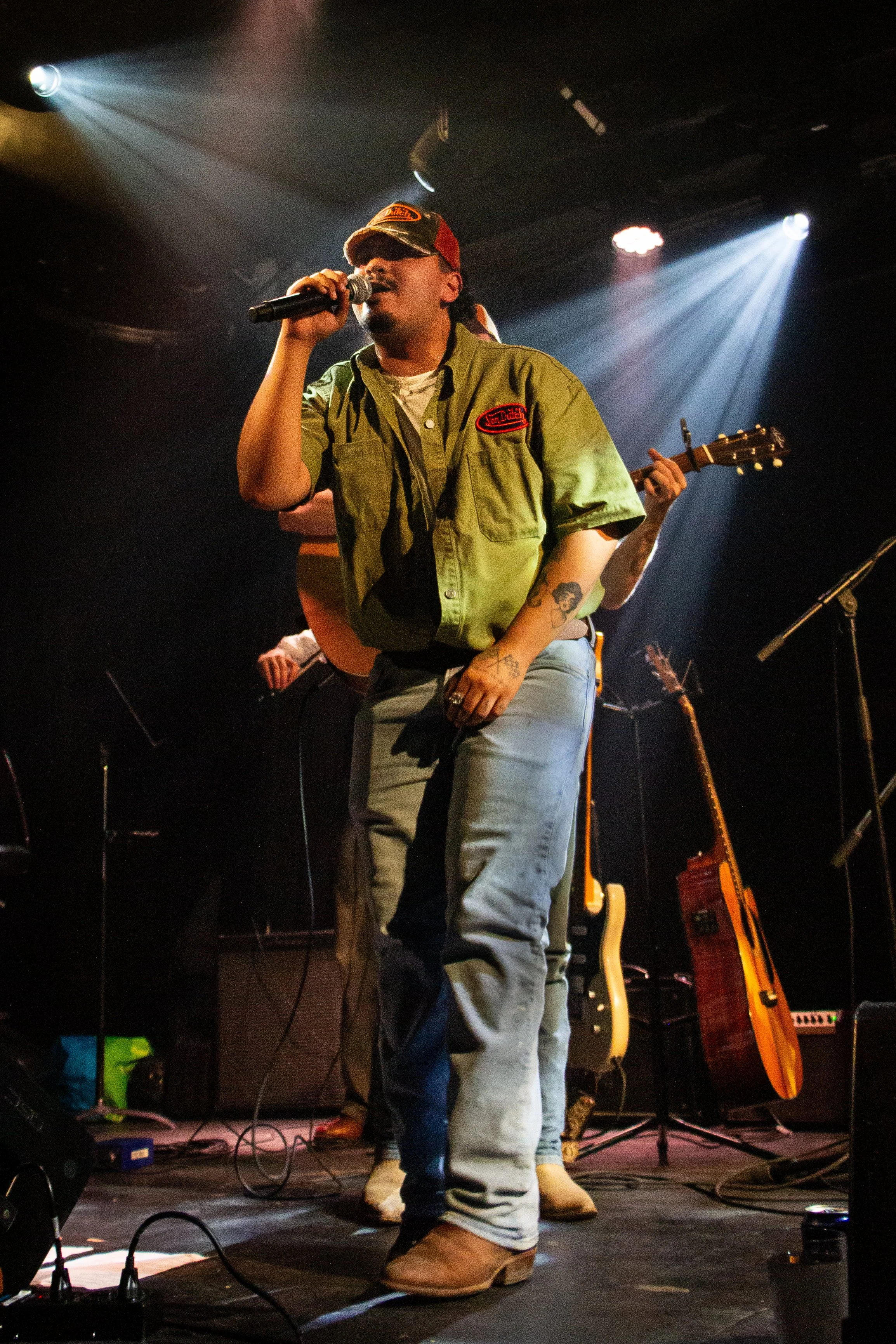 A male singer wearing a green work shirt, jeans, and cowboy boots performing on stage with a microphone, with a bearded tattooed arm. Another musician playing guitar is partially visible behind him. Stage lighting with spotlights and multiple guitars