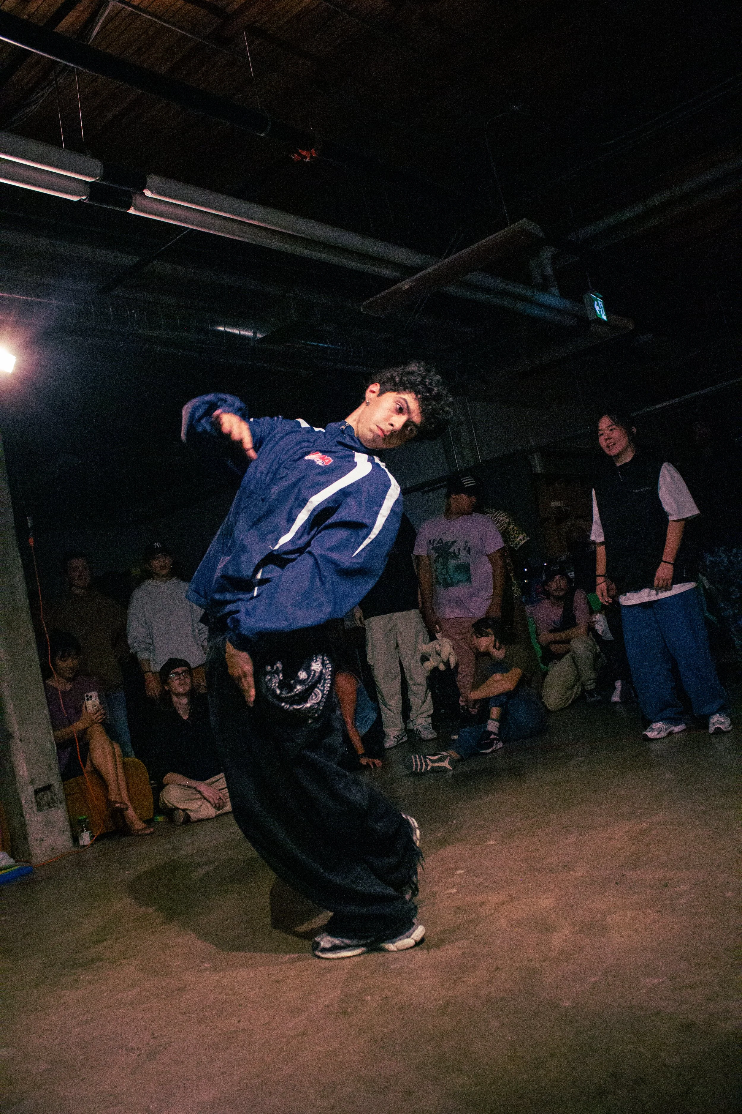 A young man dancing in a dimly lit indoor space with a crowd of people watching him, some taking photos.