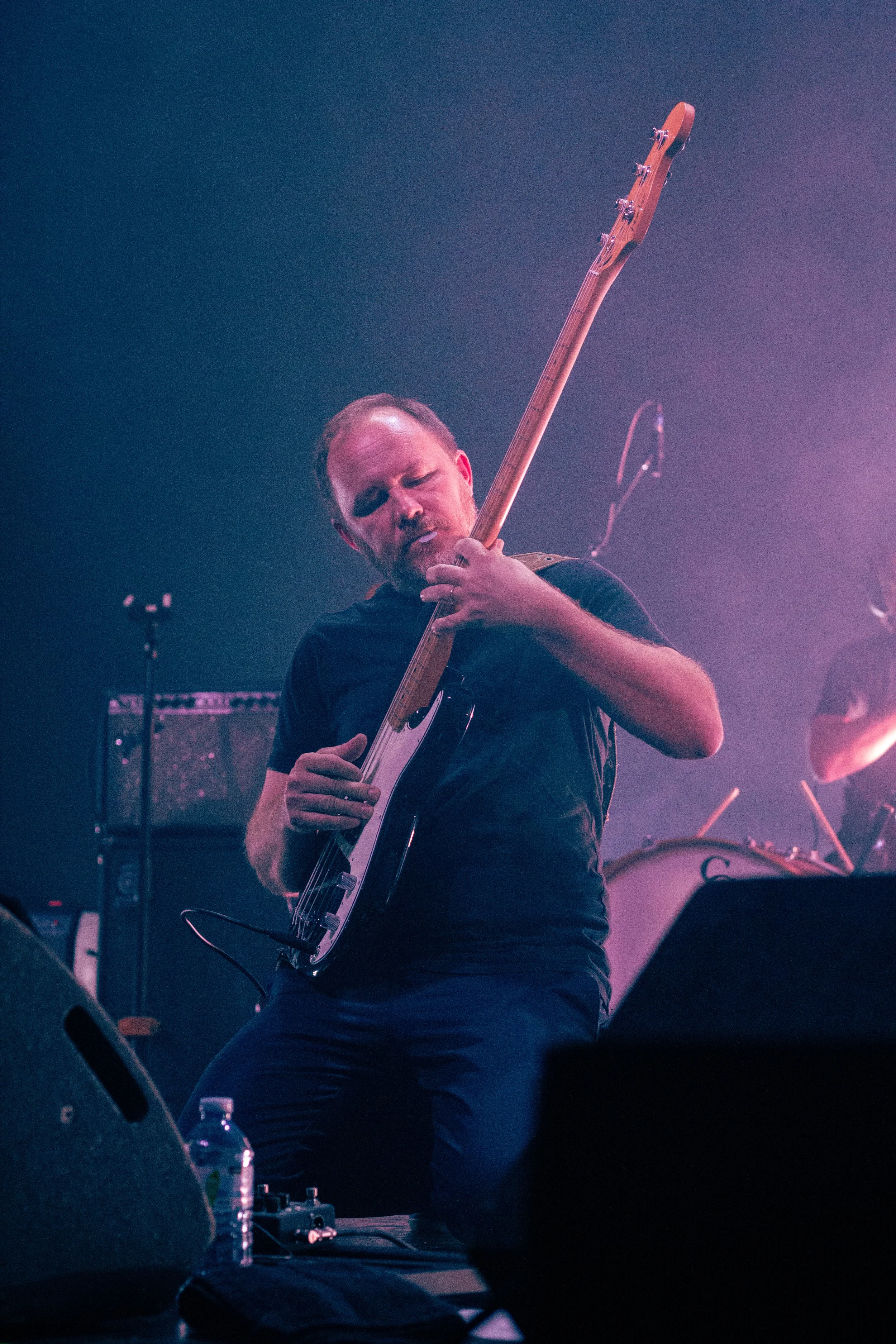 A man with a beard and short hair is playing an electric guitar on stage with purple and blue lighting. He is kneeling and intensely focused on his performance.
