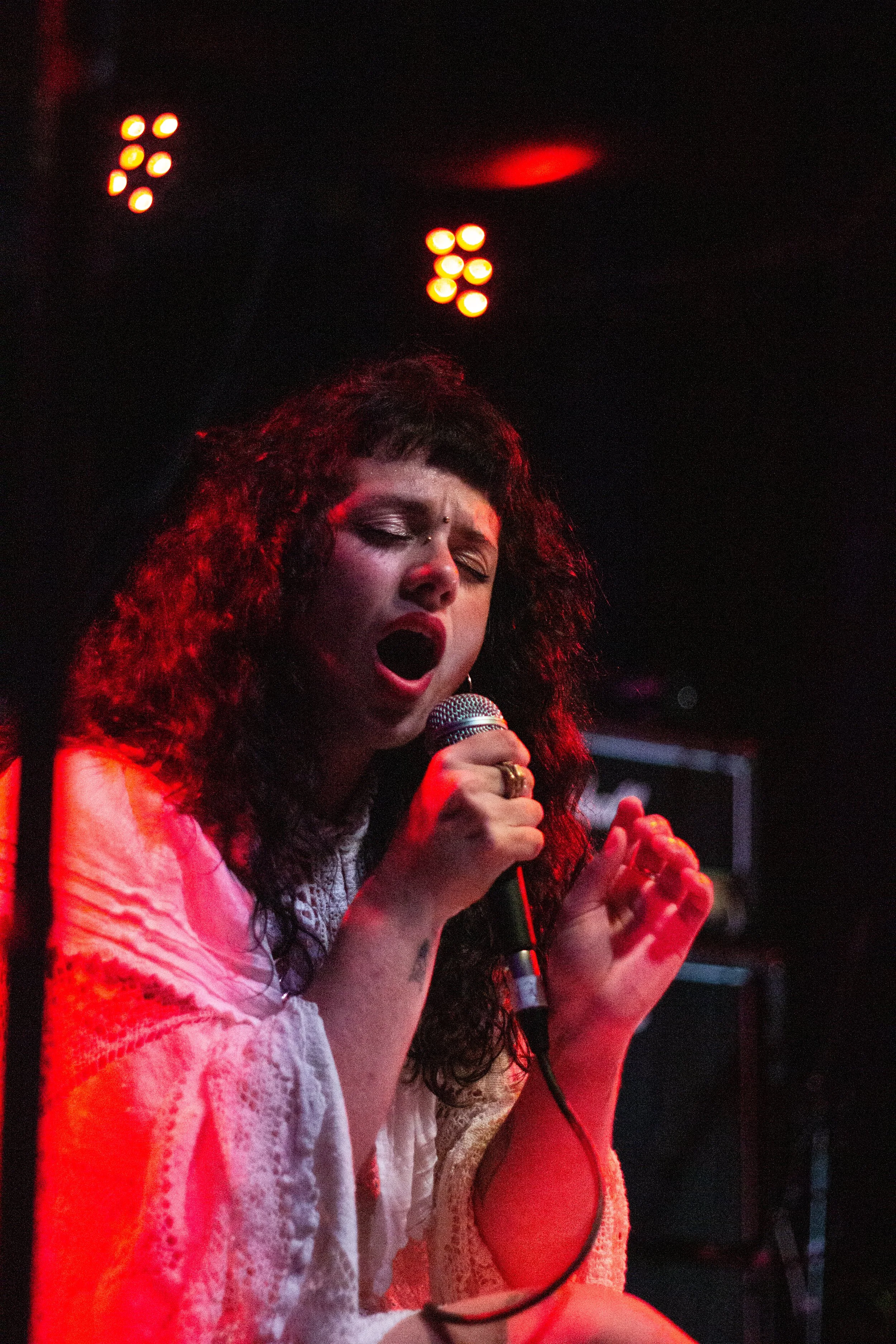 A woman with curly dark hair and a small bindi on her forehead, singing into a microphone on stage in a dimly lit environment with red stage lighting.