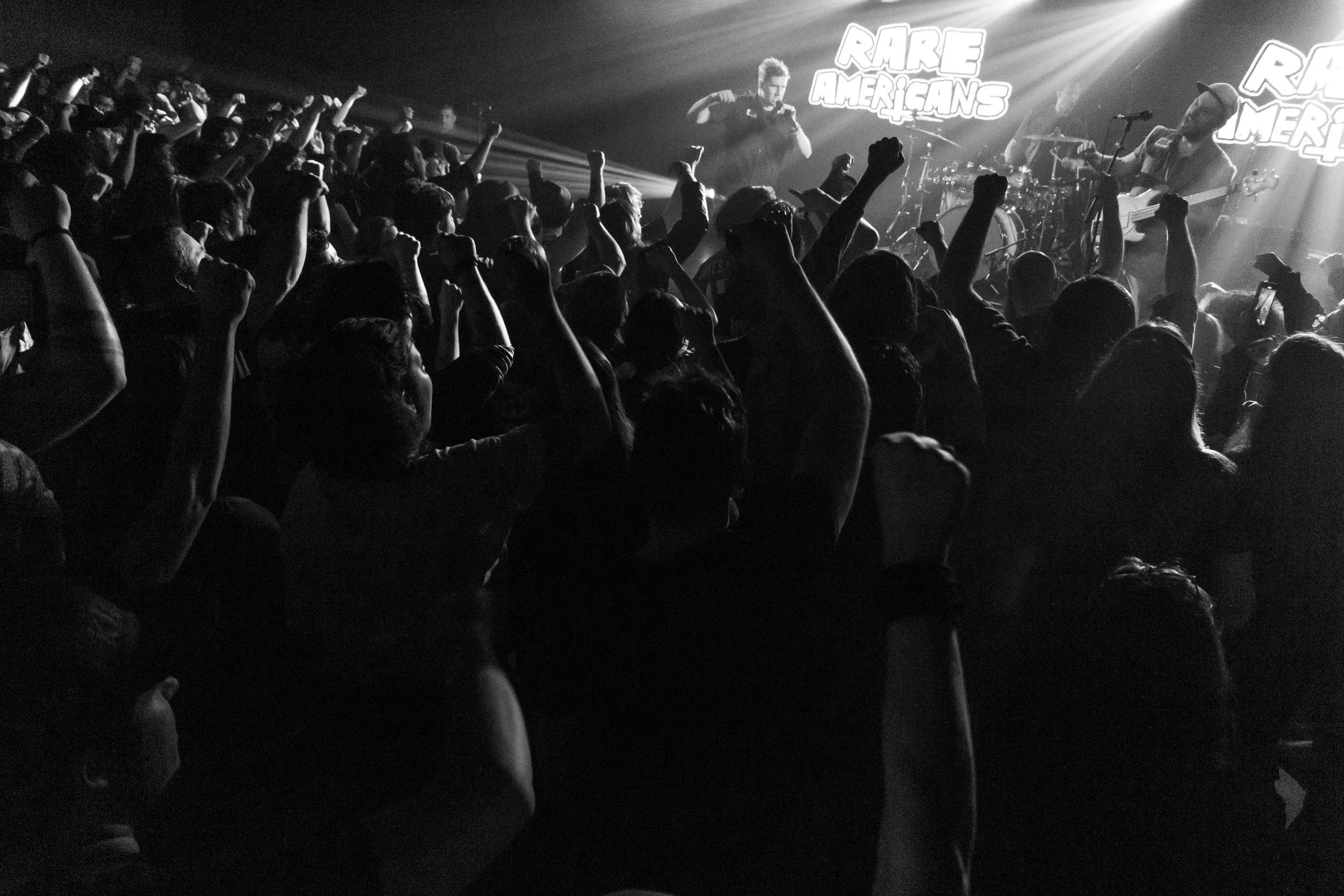 A black and white photo of a concert with a crowd of people raising their fists, facing a stage with musicians performing. The stage has a sign that says 'RARE AMERICANS.' The audience appears engaged and energetic.