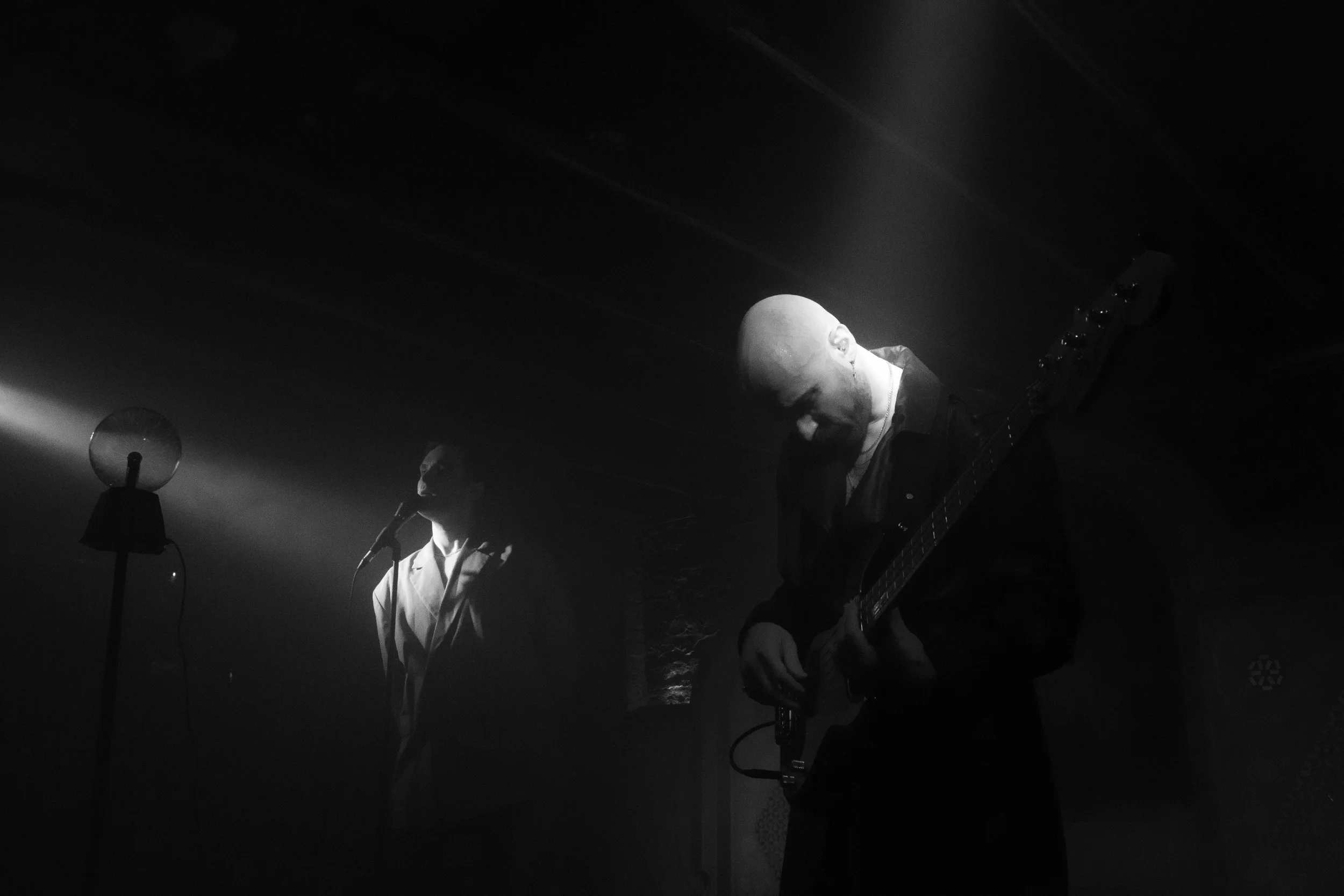 Two men performing on stage in a dark setting, one playing a guitar and the other singing into a microphone, with a microphone stand visible.