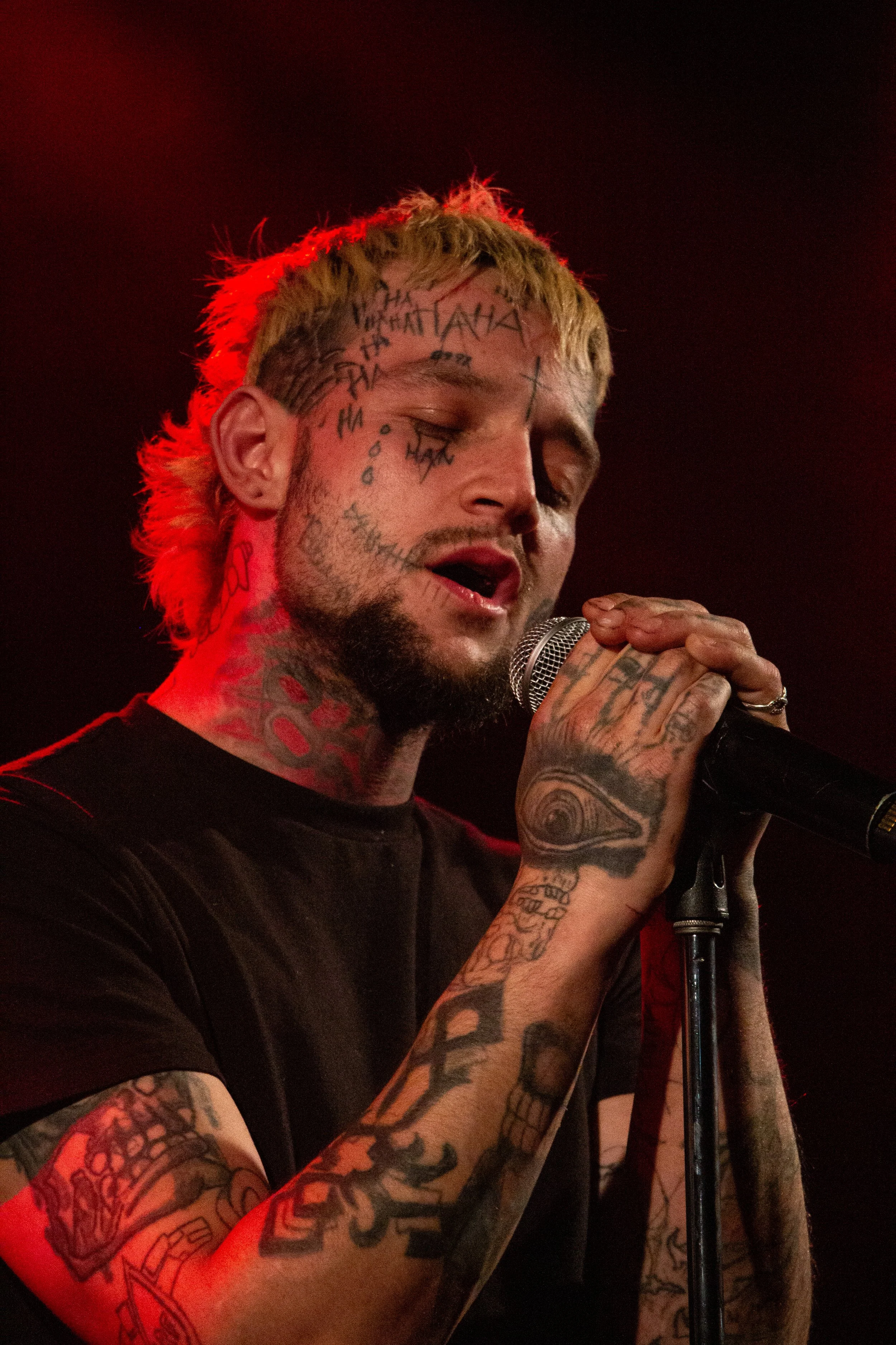 Close-up of a tattooed man singing into a microphone with eyes closed, background dark, red lighting highlighting his hair and tattoos.