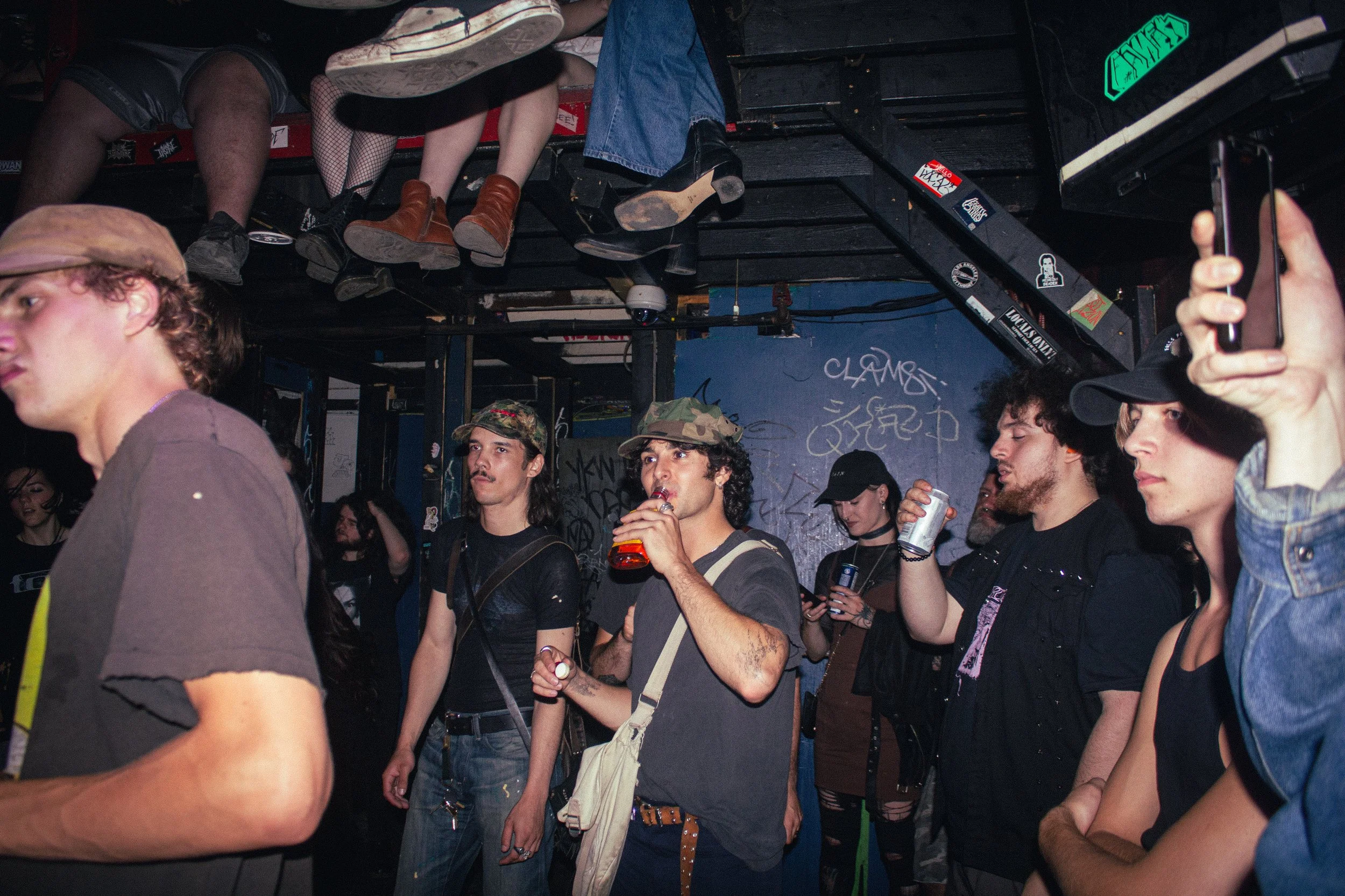 A group of people socializing at a nightclub or bar, with several people sitting and standing, some drinking, and a man in the foreground wearing a camouflage hat. The environment has graffiti on the walls and a black ceiling with exposed pipes and e