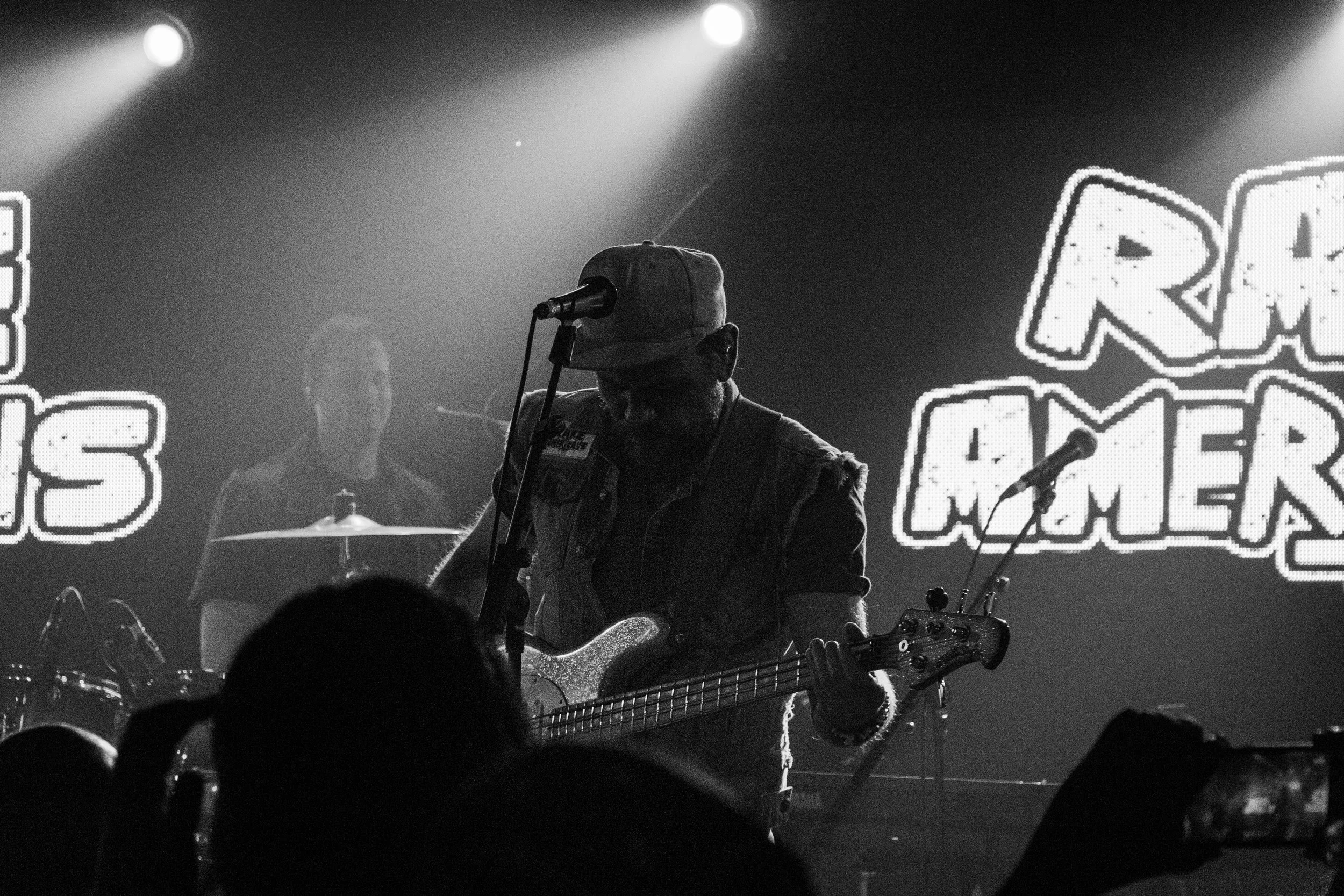 A musician playing an electric guitar on stage during a concert, with a microphone in front of him and a large illuminated sign in the background.