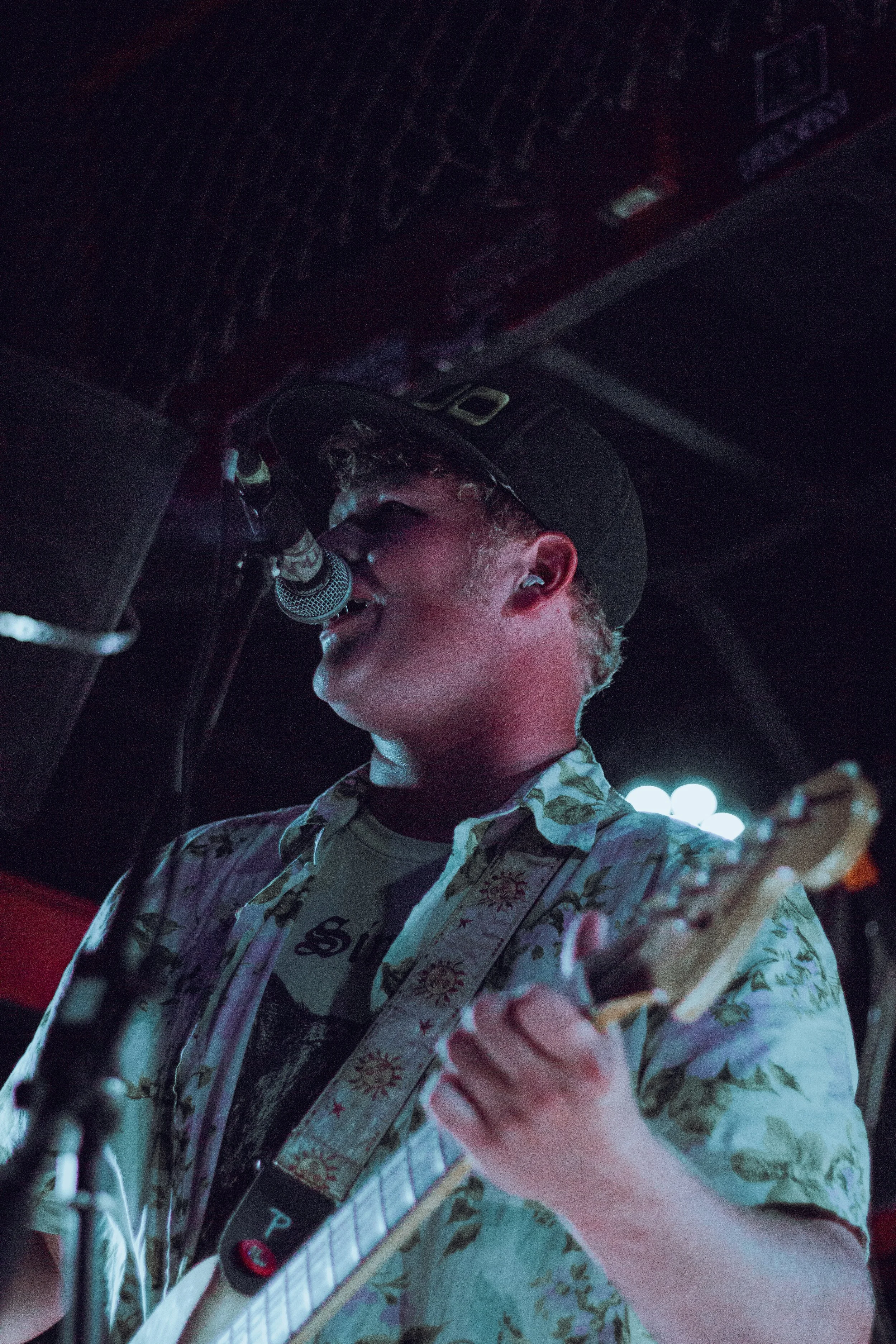 A person wearing a gray baseball cap and a floral shirt, singing into a microphone and playing an electric guitar in a dimly lit setting.