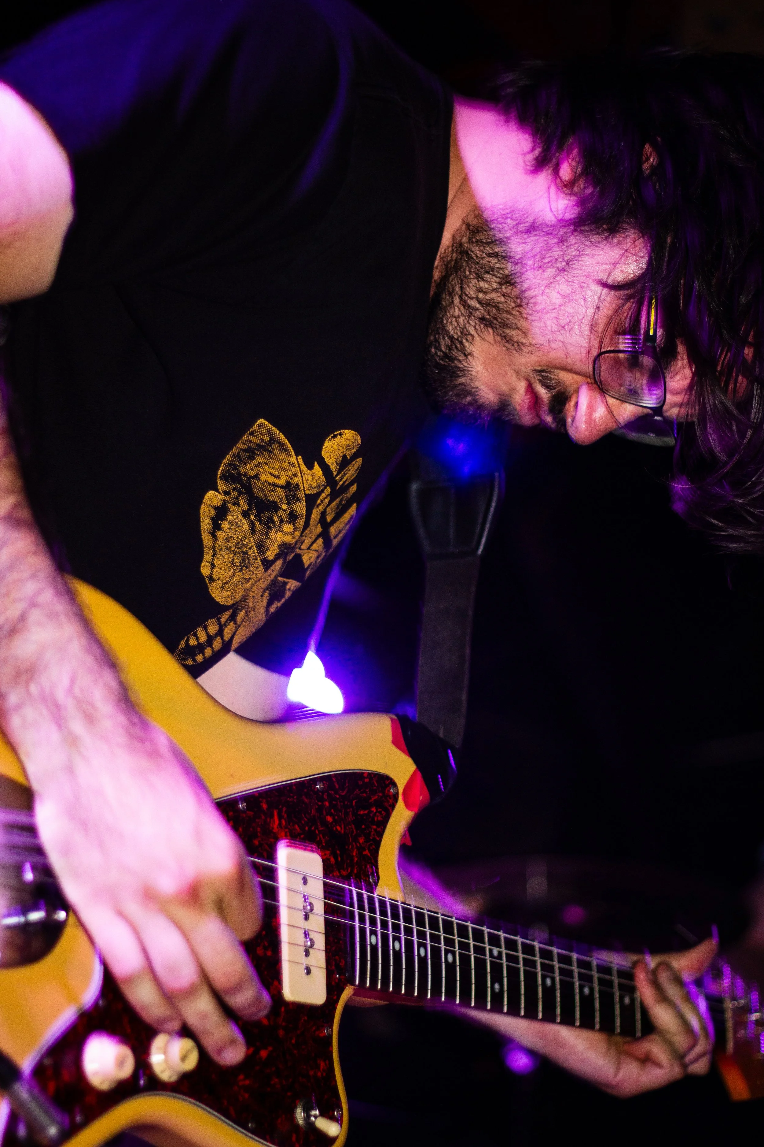 A musician playing an electric guitar with a yellow body and a red and black pickguard under purple stage lighting.
