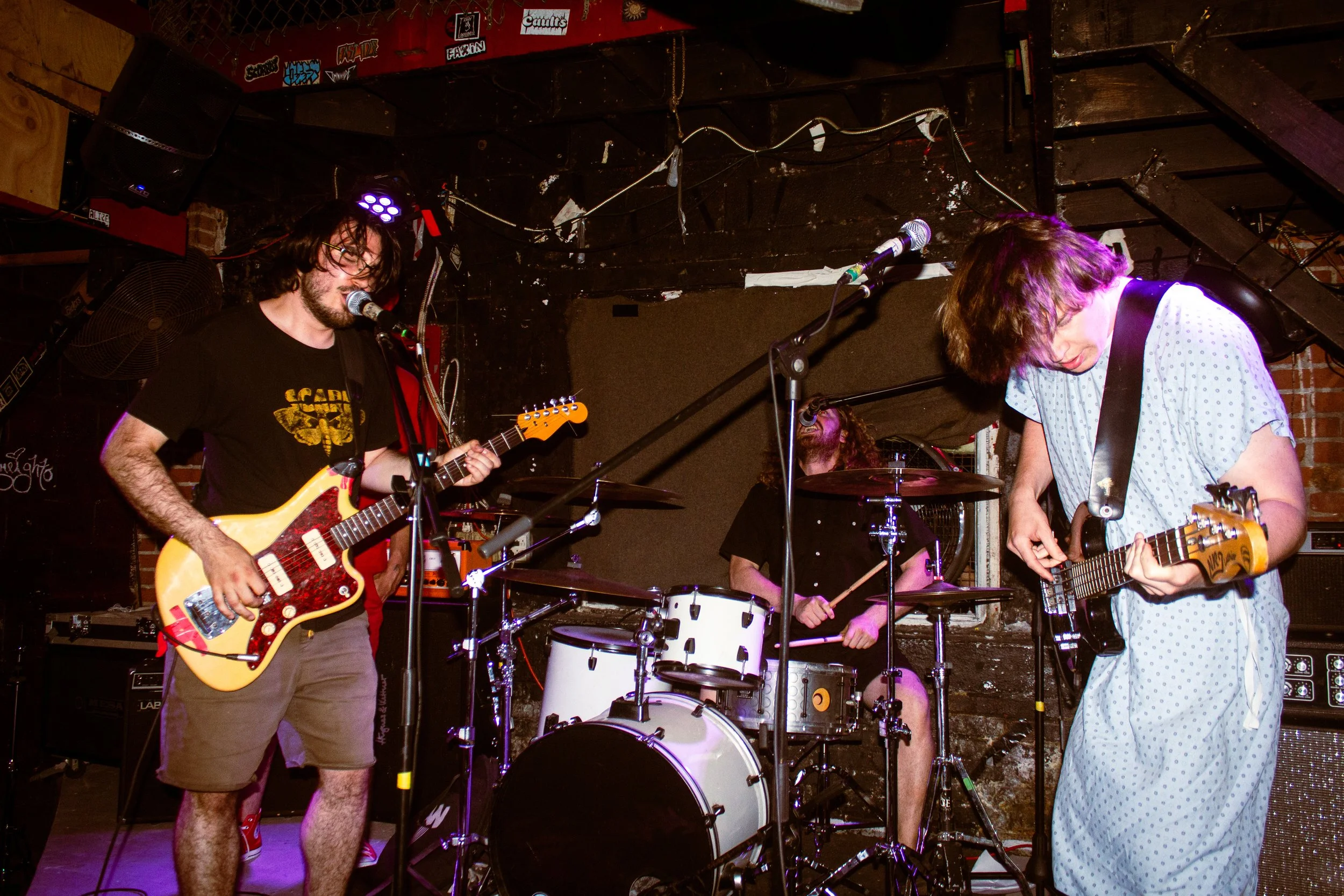 A band performing on stage with a guitarist singing into a microphone, another guitarist playing, a drummer behind, and colorful stage lighting.